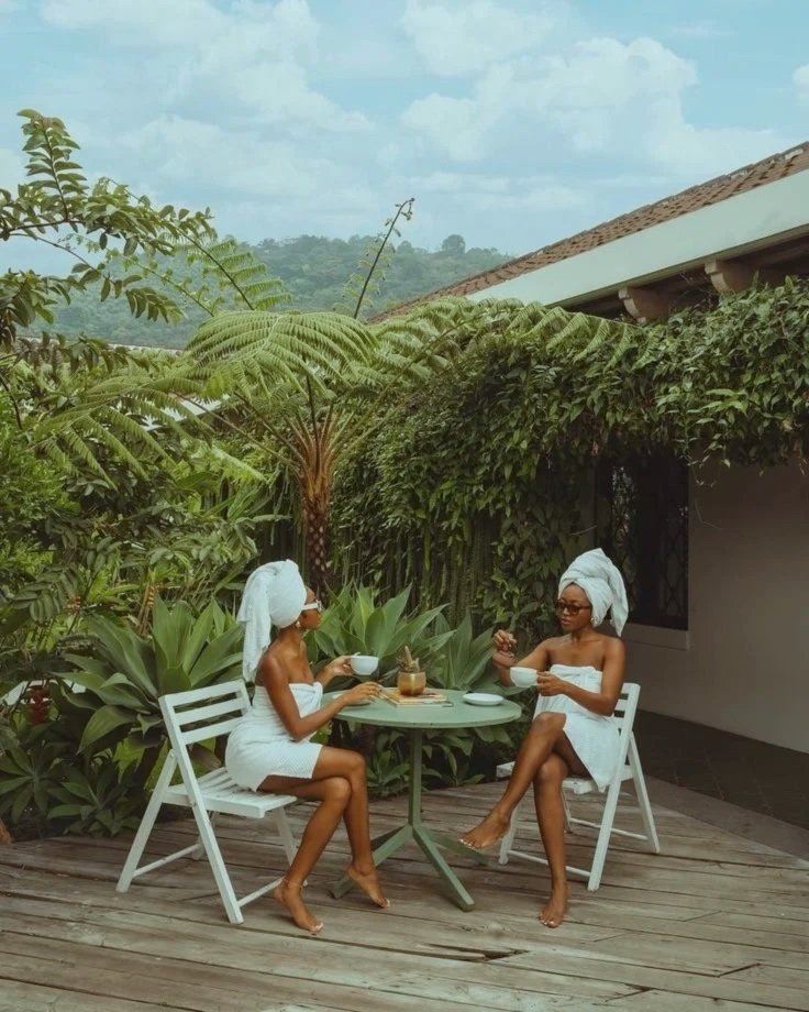 Dos mujeres con toallas y turbantes en la cabeza, tomando café en una terraza rodeada de vegetación tropical.