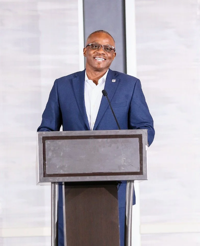 A man in a blue suit and glasses standing at a podium and smiling.
