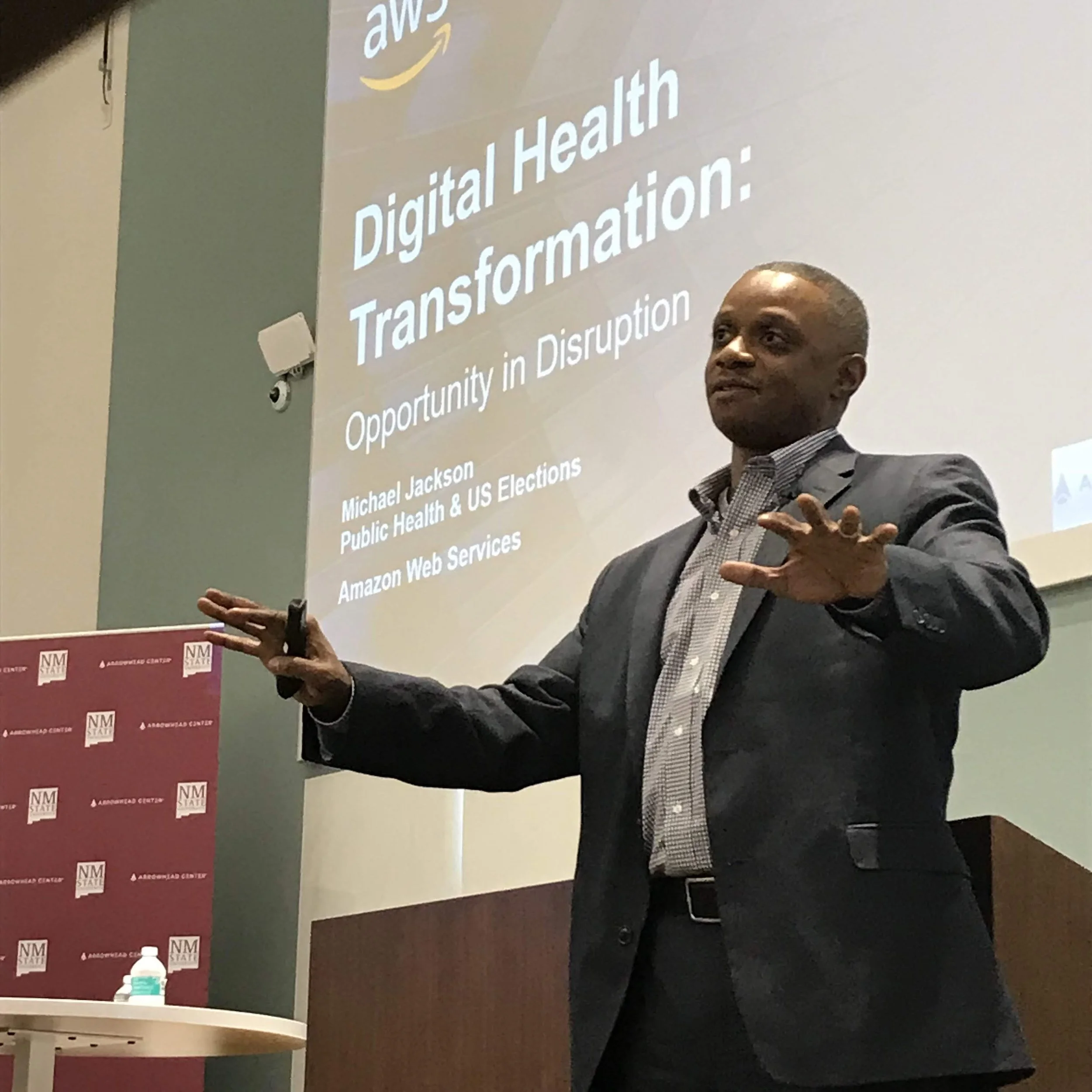 A man in a suit and checkered shirt is giving a presentation in front of a large screen. The screen displays text about digital health transformation and opportunities in the sector, with logos for NM State and Albuquerque Center.