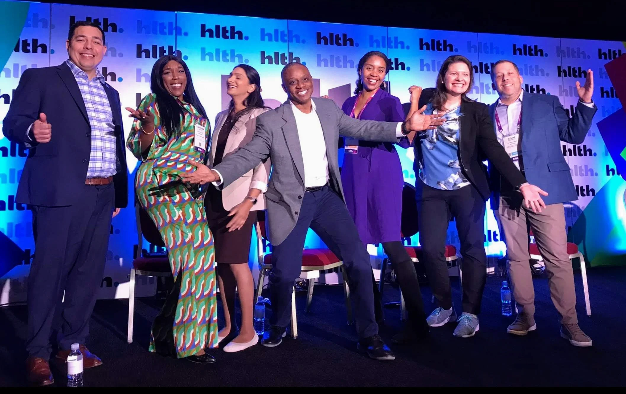 Group of seven diverse people standing on stage, smiling and making gestures, in front of a backdrop with 'hlth' repeated multiple times, during a conference or event.