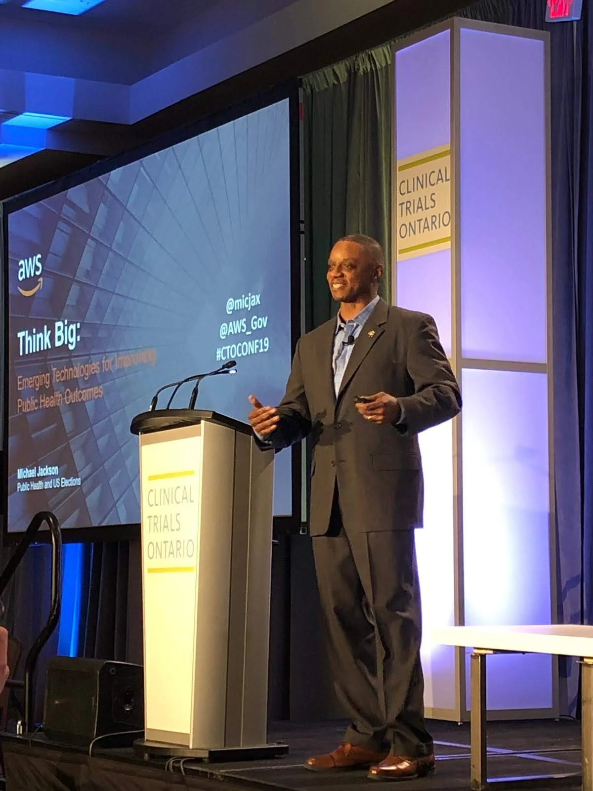 A man in a suit speaking at a podium during a conference in front of a screen that displays a presentation slide. The slide reads 'Think Big: Emerging Technologies for Improving Public Health Outcomes' with social media handles and hashtag, and the conference is associated with Clinical Trials Ontario.