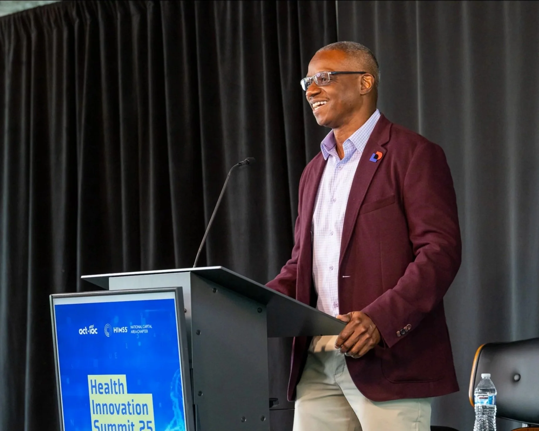 A man stands at a podium giving a speech or presentation at an event, with a dark curtain as the backdrop. He wears glasses, a maroon blazer with a pin, a light-colored shirt, and beige pants. There is a water bottle on the table beside him, and a monitor displaying a blue screen with yellow text related to health, innovation, and summits.