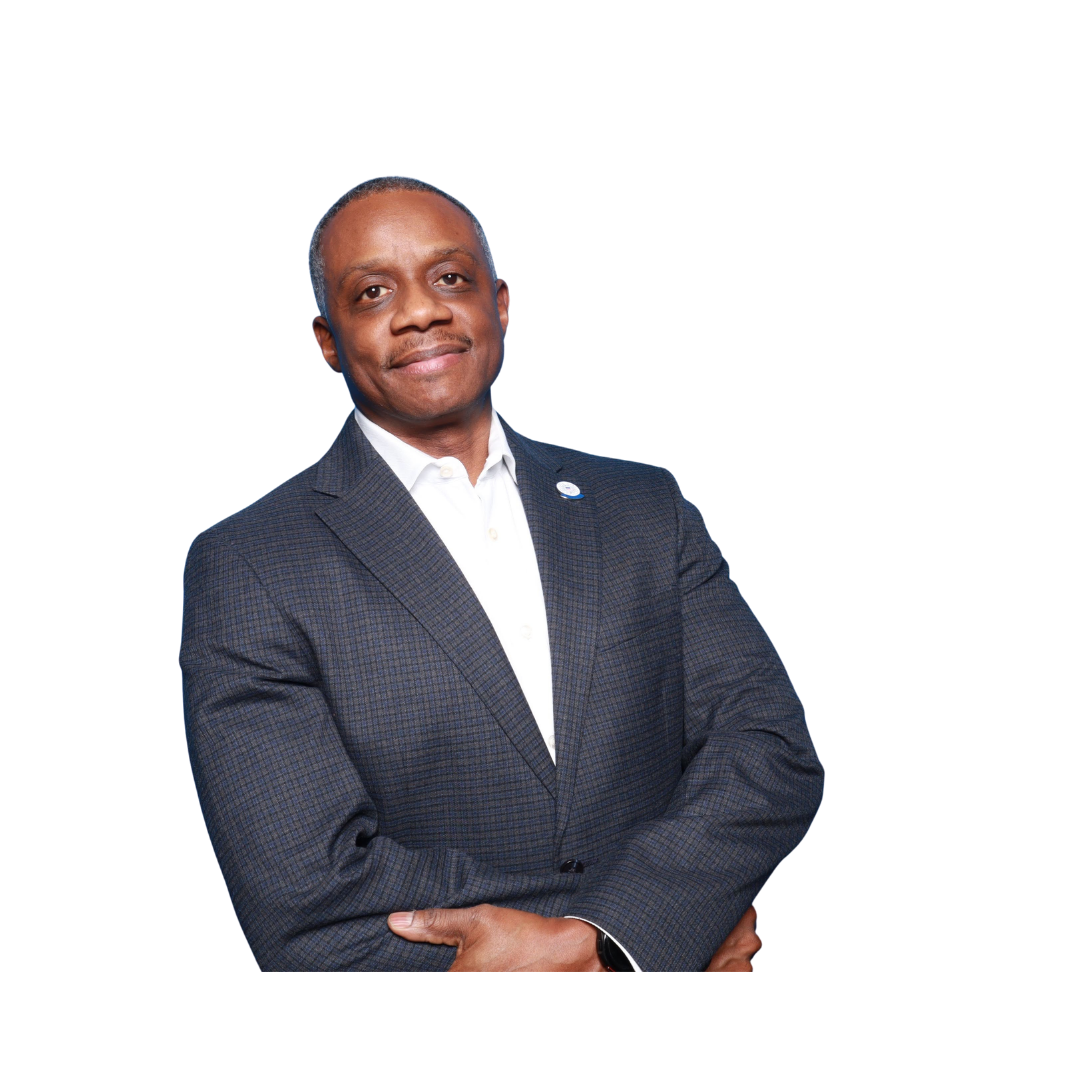A middle-aged African American man in a dark checkered suit and white shirt, standing with arms crossed, smiling confidently against a plain dark background.