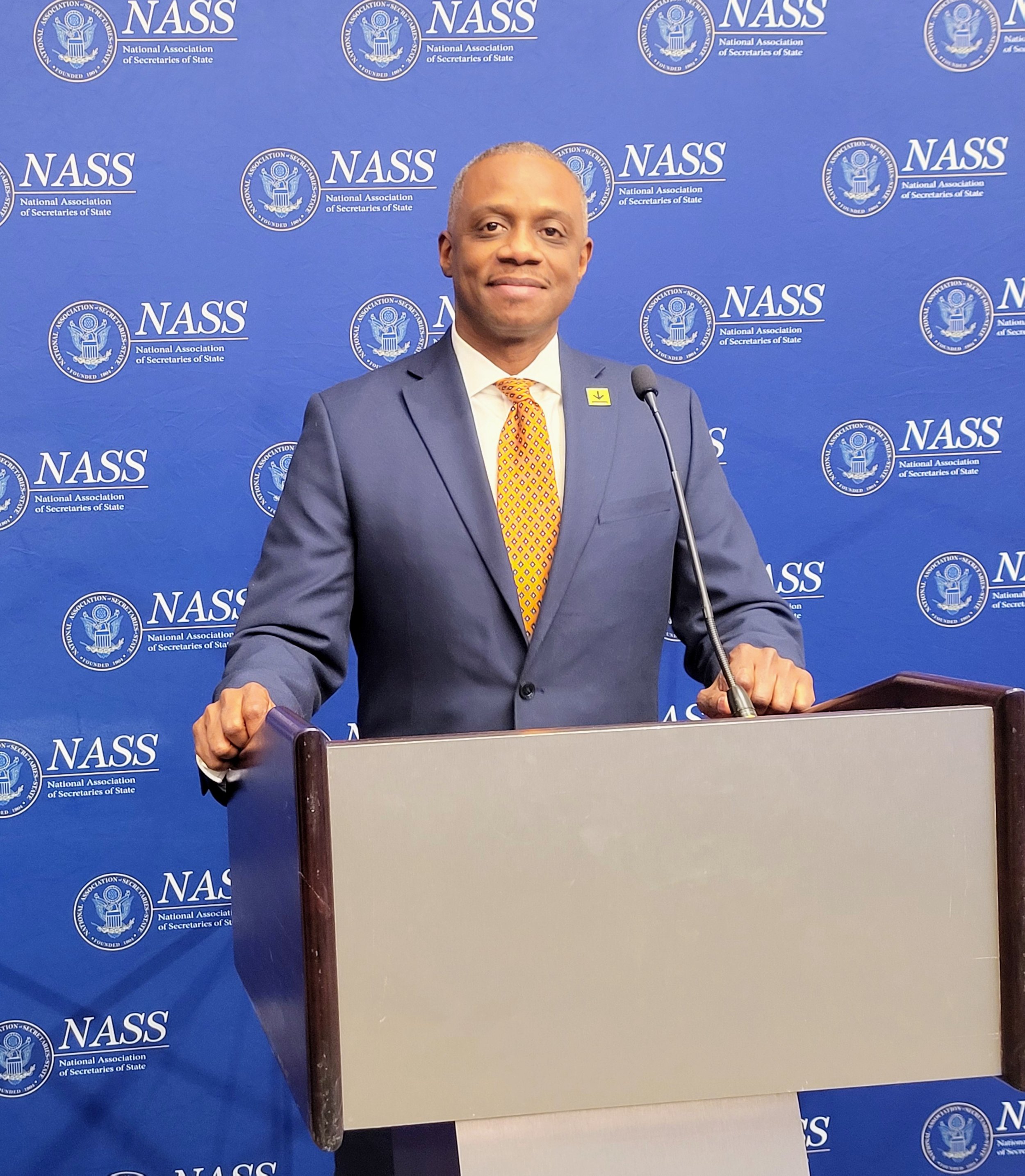 A man in a dark suit and tie standing behind a podium at a press conference, with a blue backdrop featuring the logo and name of the National Association of Secretaries of State (NASS).
