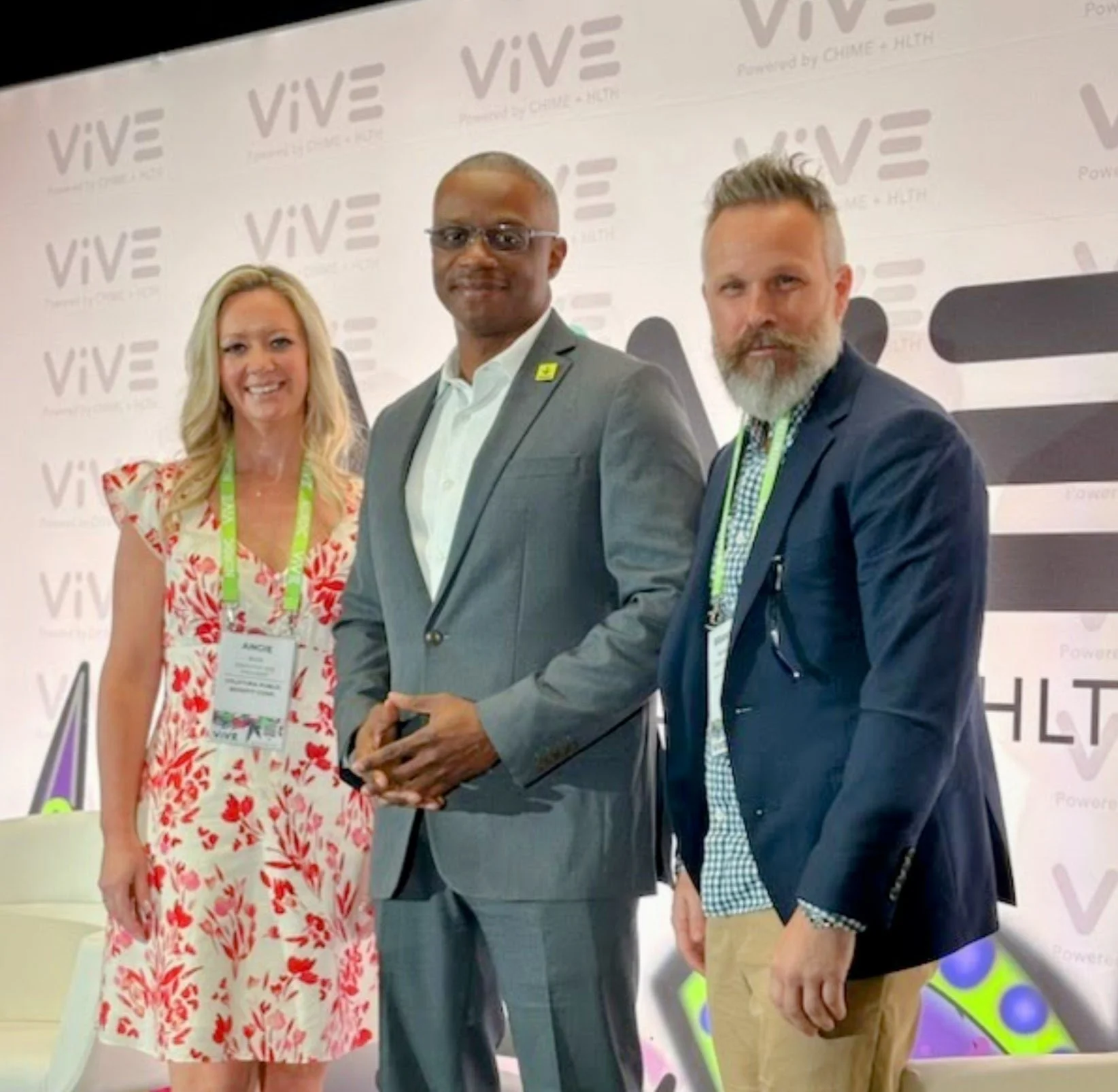 Three professionally dressed people standing together at a conference or event with a background that reads 'VIVE' and 'Power of CIME + HEH'.