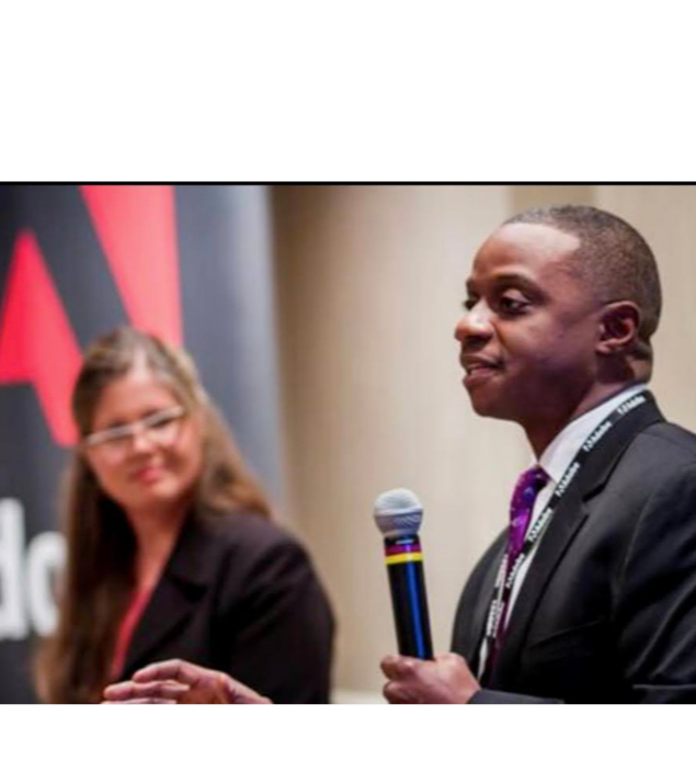 A man in a suit holding a microphone, speaking at an event, with a blurred woman in glasses listening in the background.