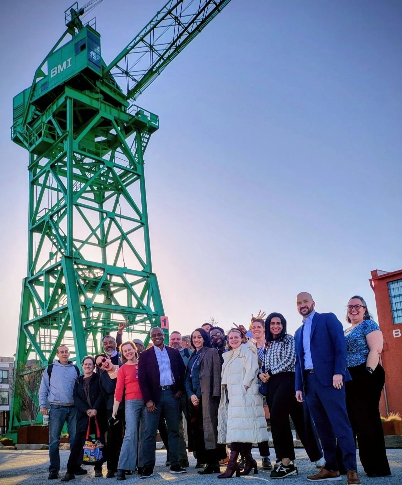 A diverse group of people standing together outdoors, smiling, with a large green crane structure labeled 'BM1' in the background, and buildings around as the sky is clear.