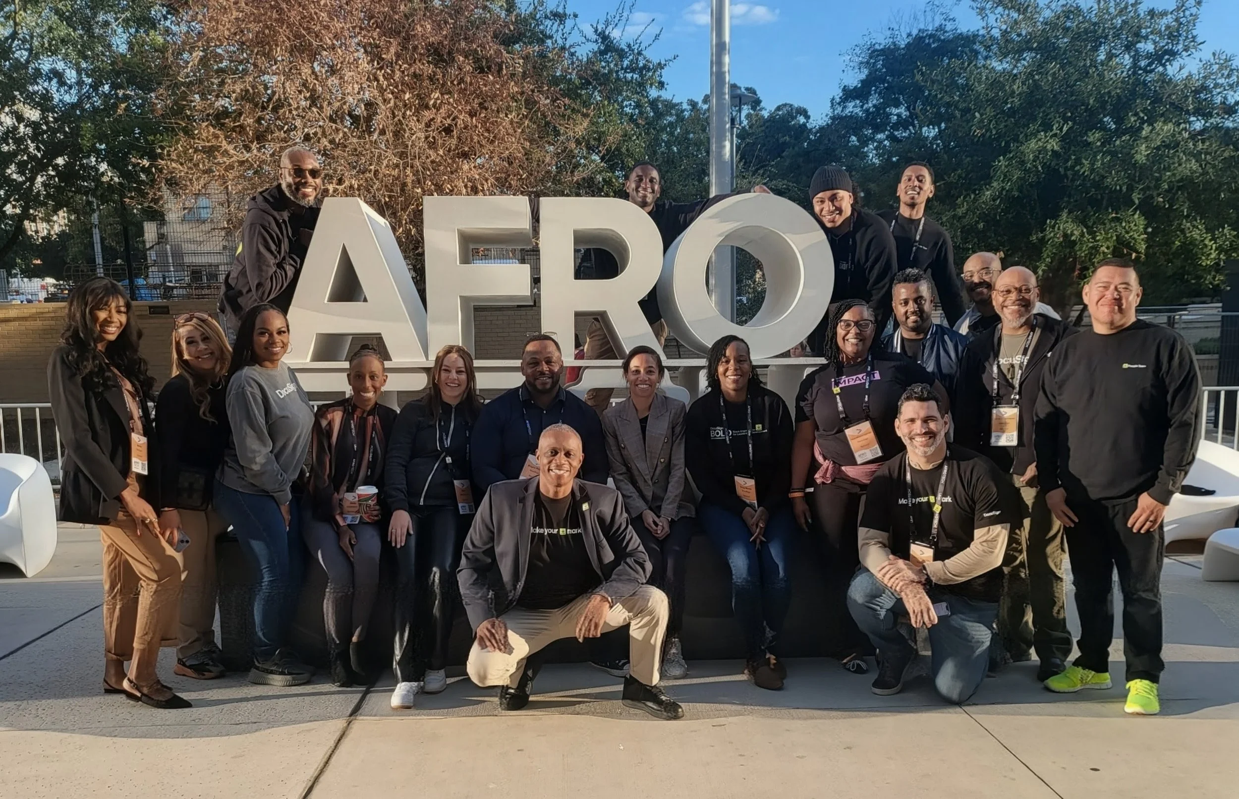 A group of people outdoors, smiling, posing for a photo in front of large white letters spelling 'AFRO' with trees and a clear blue sky in the background.