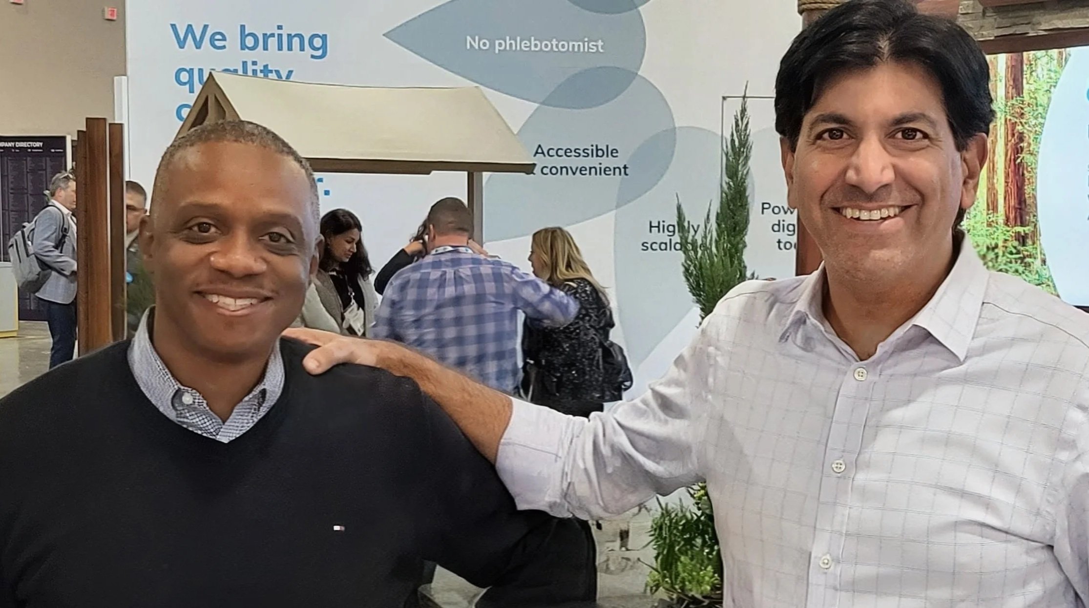 Two men smiling and posing for a photo at a conference or event, with a sign in the background that reads, "We bring quality," and a group of people talking. One man has short black hair and is wearing a black sweater over a collared shirt, while the other has dark hair and is wearing a light-colored checkered shirt.