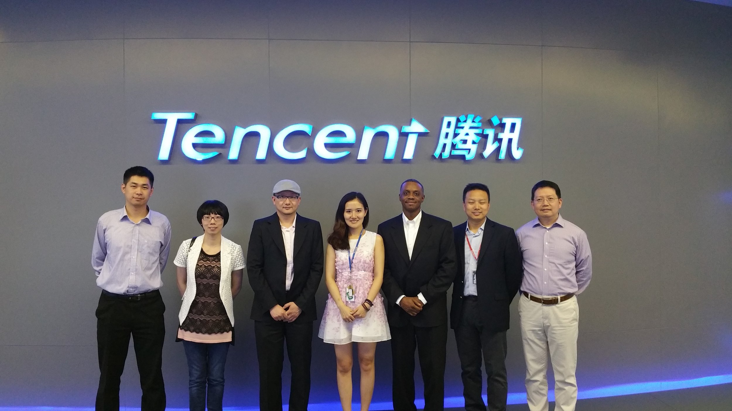 A group of seven diverse people standing in front of a wall with a large illuminated Tencent logo and Chinese characters. They are dressed in business casual and formal attire, smiling for the photo.