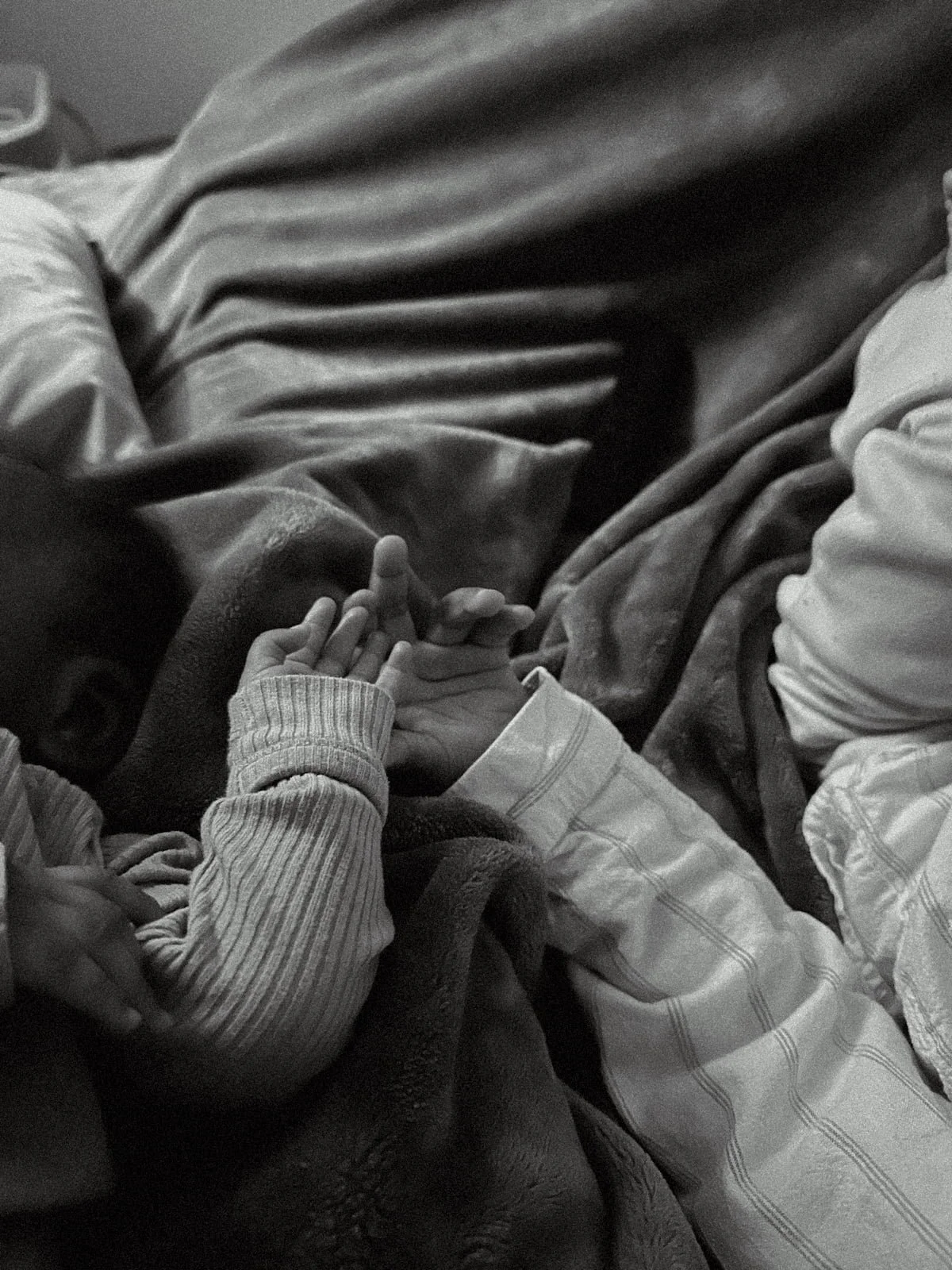 A black and white photo of a young child and an adult holding hands, lying down on blankets or soft surfaces, with a relaxed and intimate atmosphere.