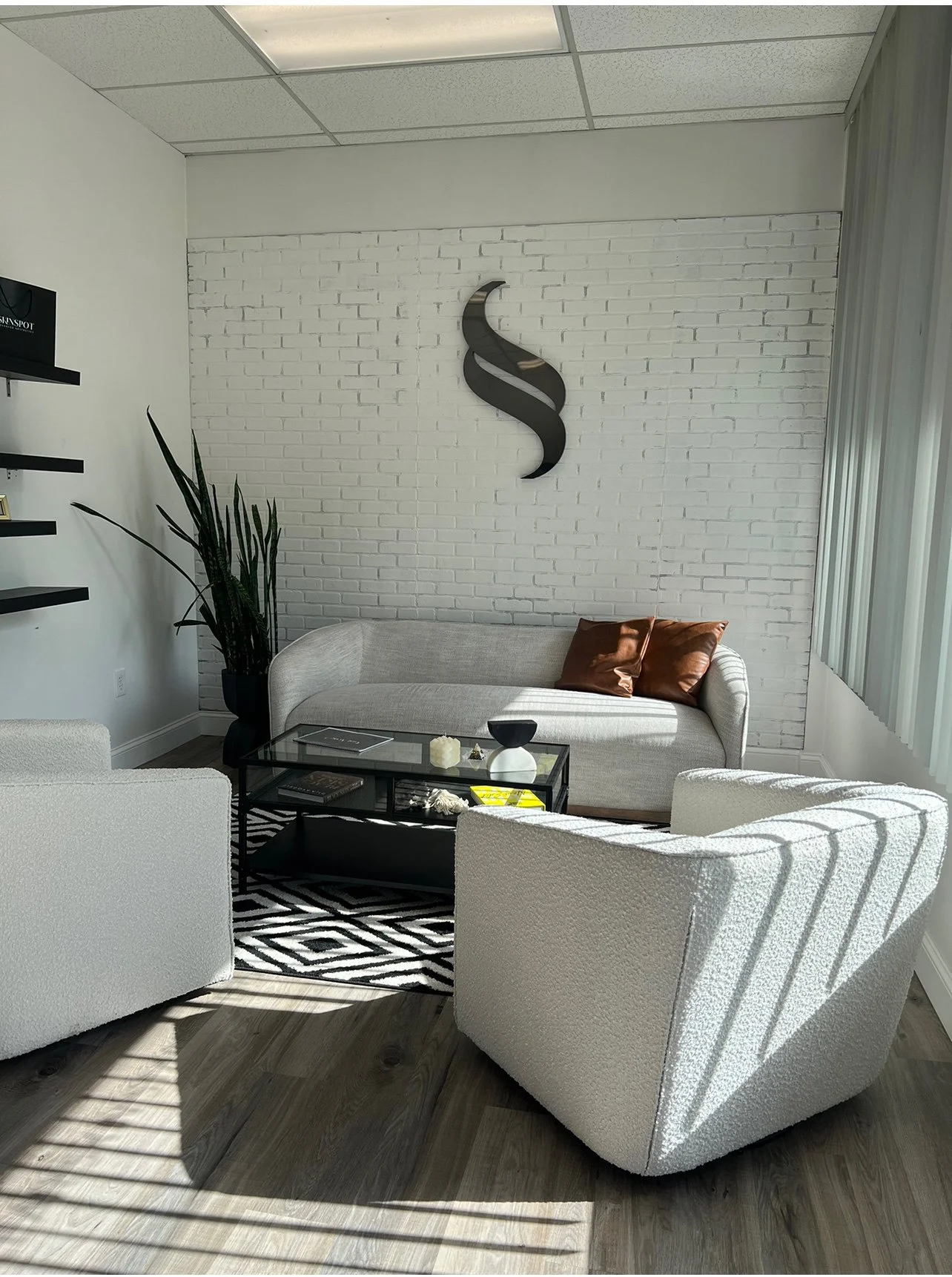 Modern waiting room with white brick accent wall, black wall art, a gray sofa with brown pillows, white armchairs, a glass coffee table with decorative items, a black and white patterned rug, indoor plants, and large window with vertical blinds letting in sunlight.