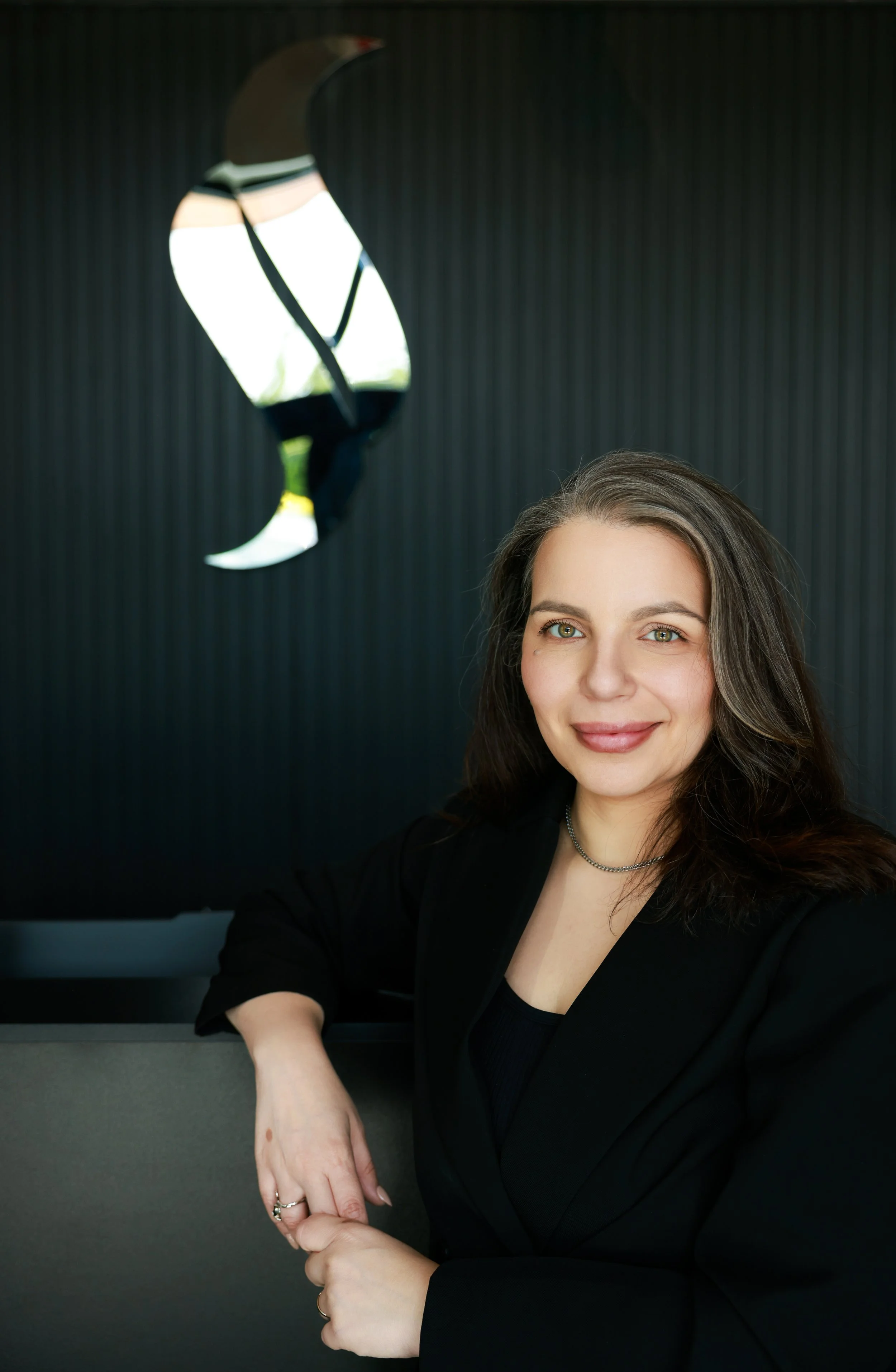 Portrait of a woman with long dark hair and blue eyes, smiling, wearing a black top and a silver necklace, sitting in front of a dark background with a reflective logo on the wall.