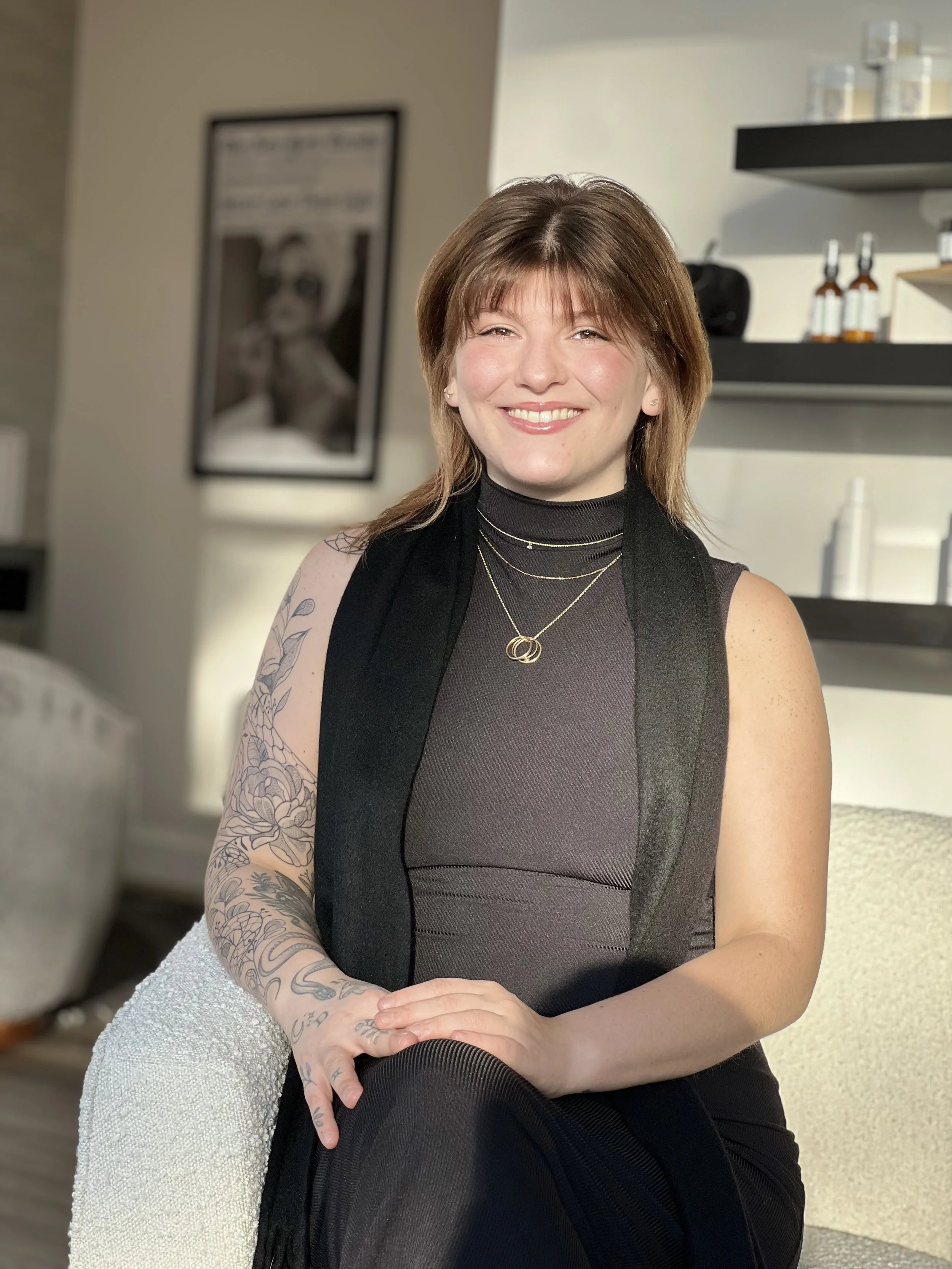 Smiling woman with brown hair, tattoos on her left arm, wearing a black sleeveless top with layered gold necklaces, sitting in a modern indoor space with shelves and a framed black-and-white photo in the background.