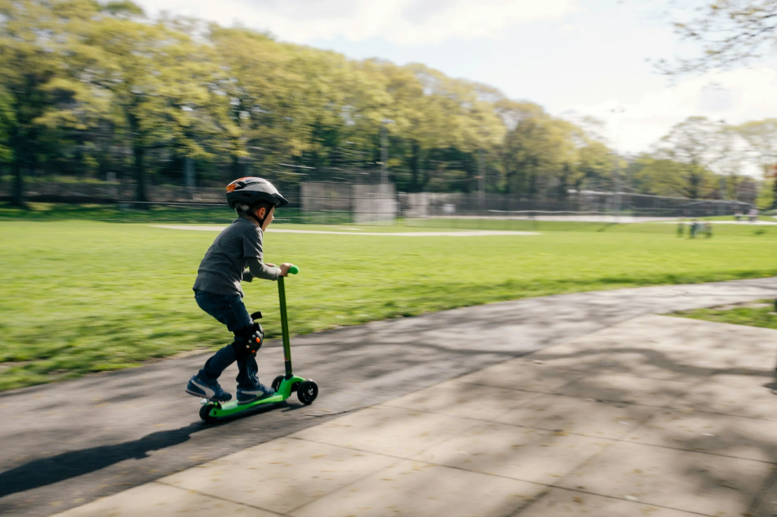 A young boy wearing a helmet riding a green scooter on a paved path in a park with green grass and trees in the background.