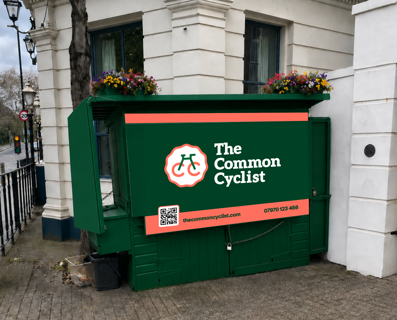 A green billboard advertising 'The Common Cyclist' with a logo of a bicycle inside a gear shape, website URL, phone number, and QR code. The billboard is situated outdoors near a building with white walls, flower boxes on top, and street lamps nearby.