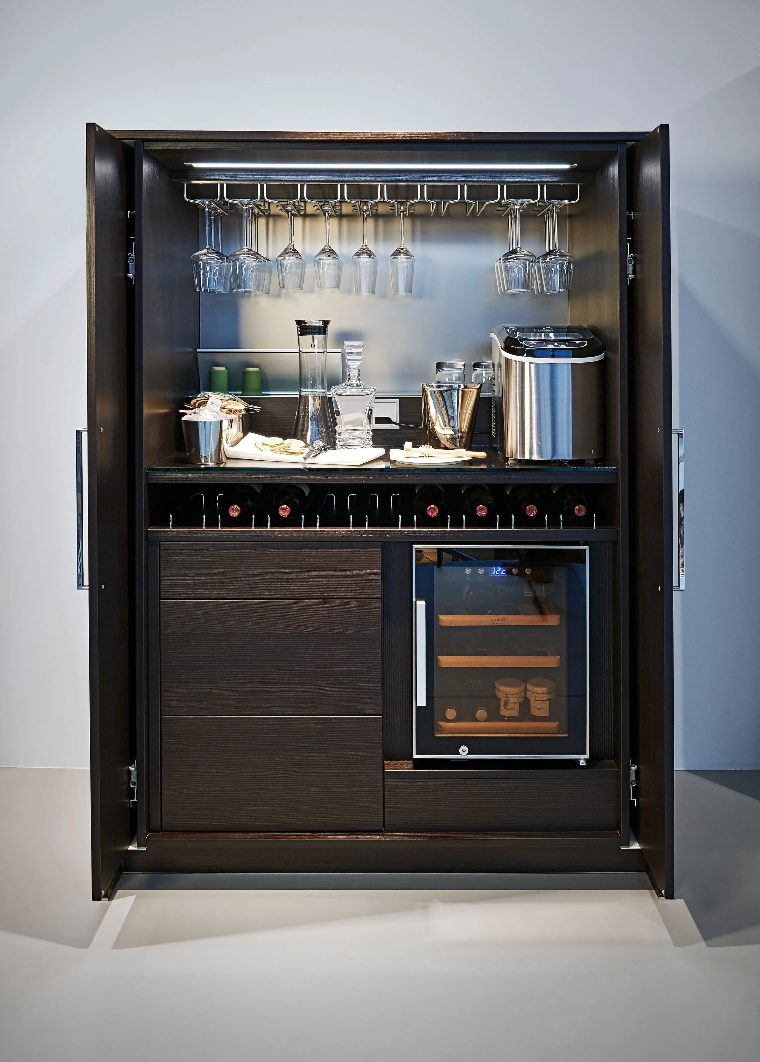 A black wooden bar cabinet with hanging wine glasses, a wine cooler, a mini fridge, and wine bottles stored in a compartment at the bottom.