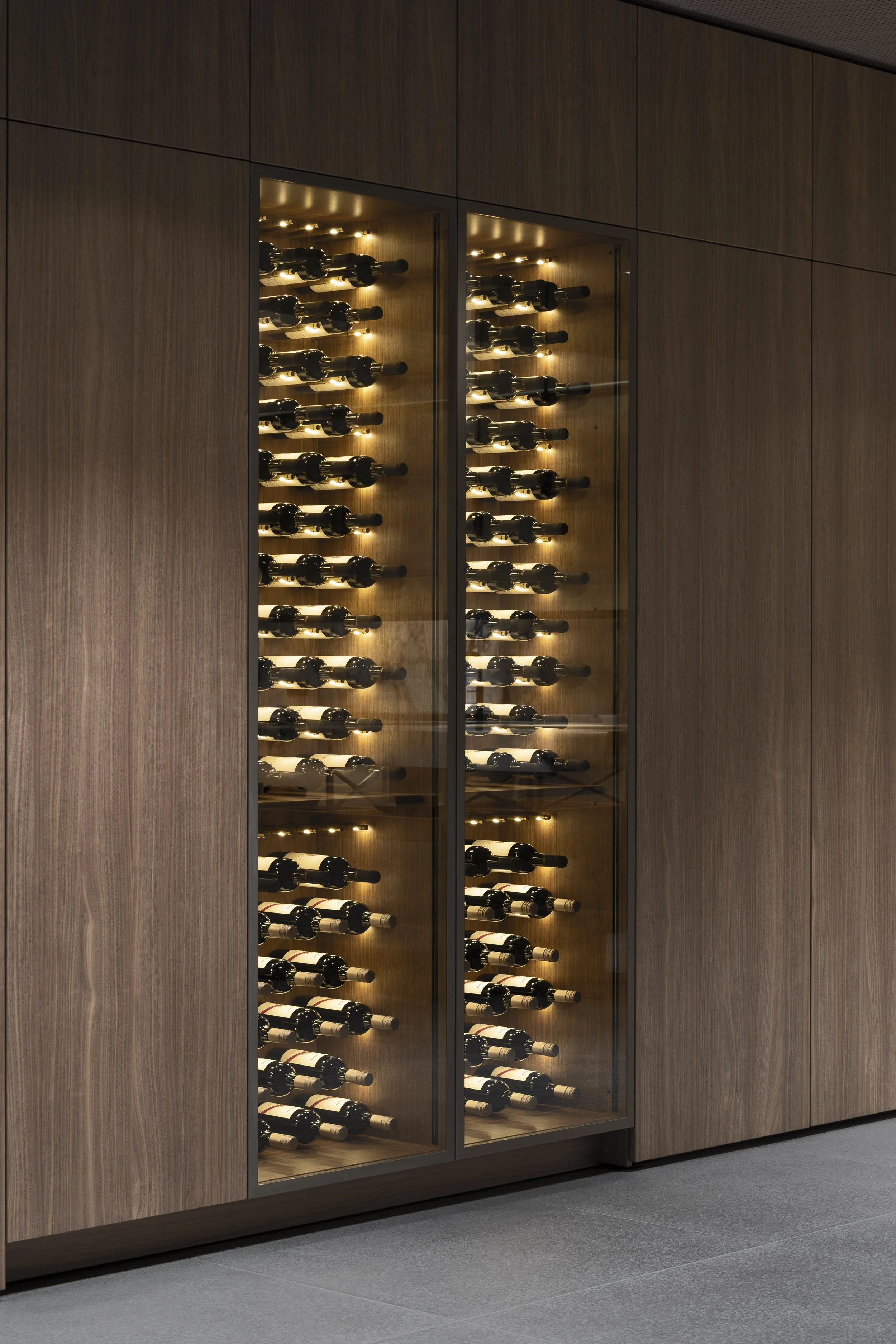 A wooden wine cellar with glass doors, illuminated shelf racks, filled with bottles of wine stored horizontally.