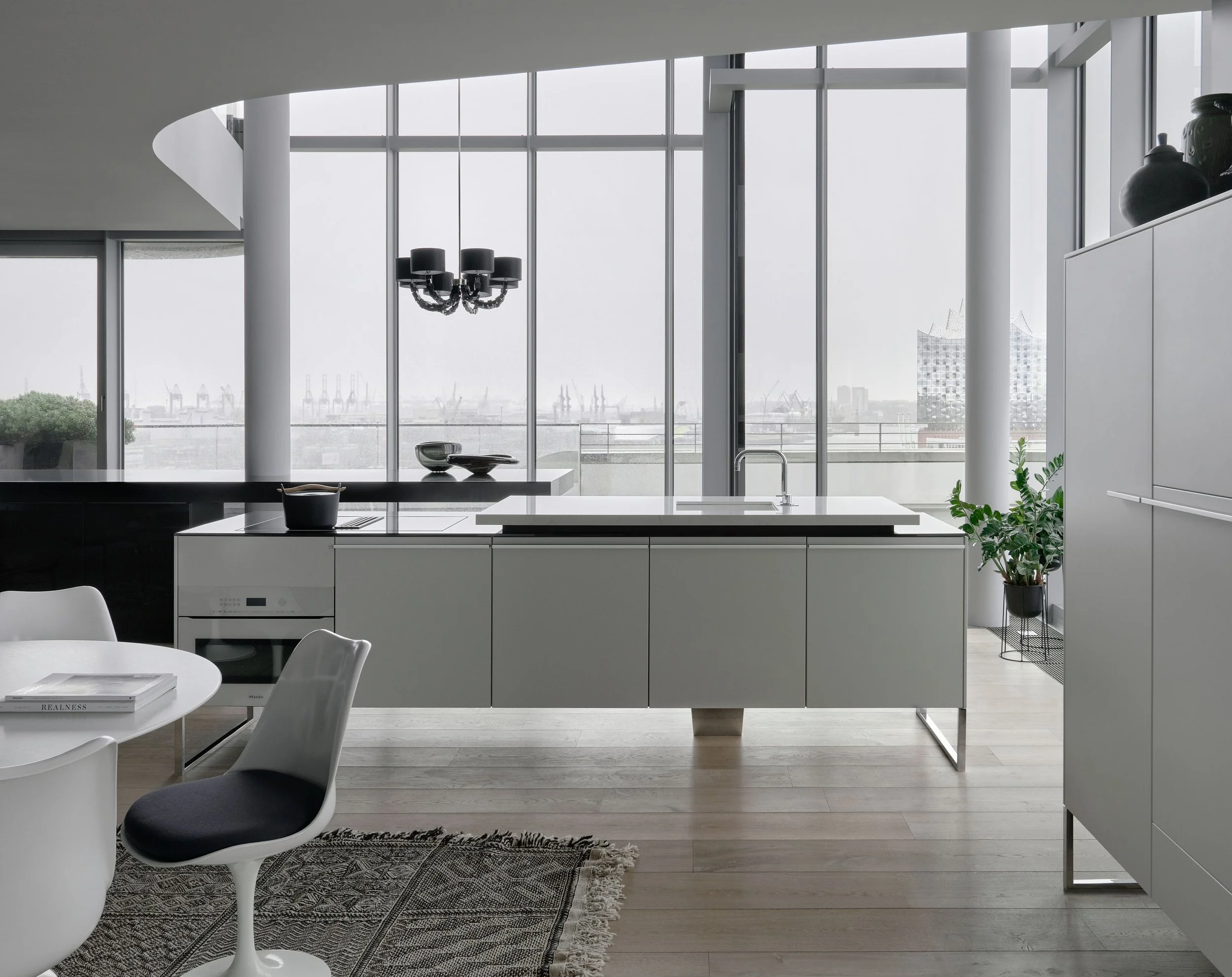 Modern kitchen with large windows showing city skyline, black chandelier, white cabinetry, kitchen island with a faucet, and a potted plant.