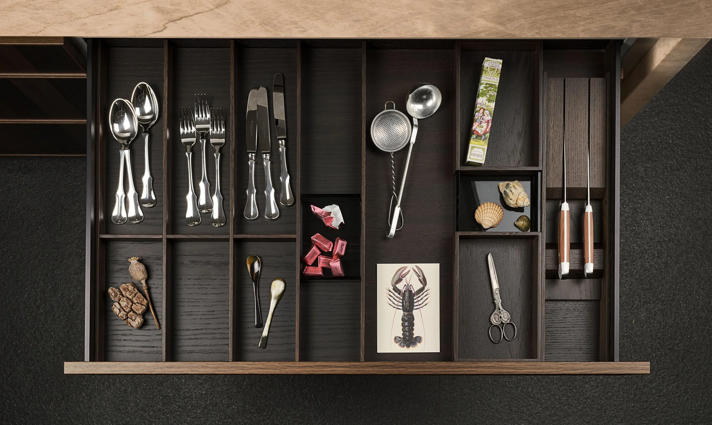 A wooden drawer divided into compartments holding silverware, shells, a lobster drawing, and miscellaneous small items.