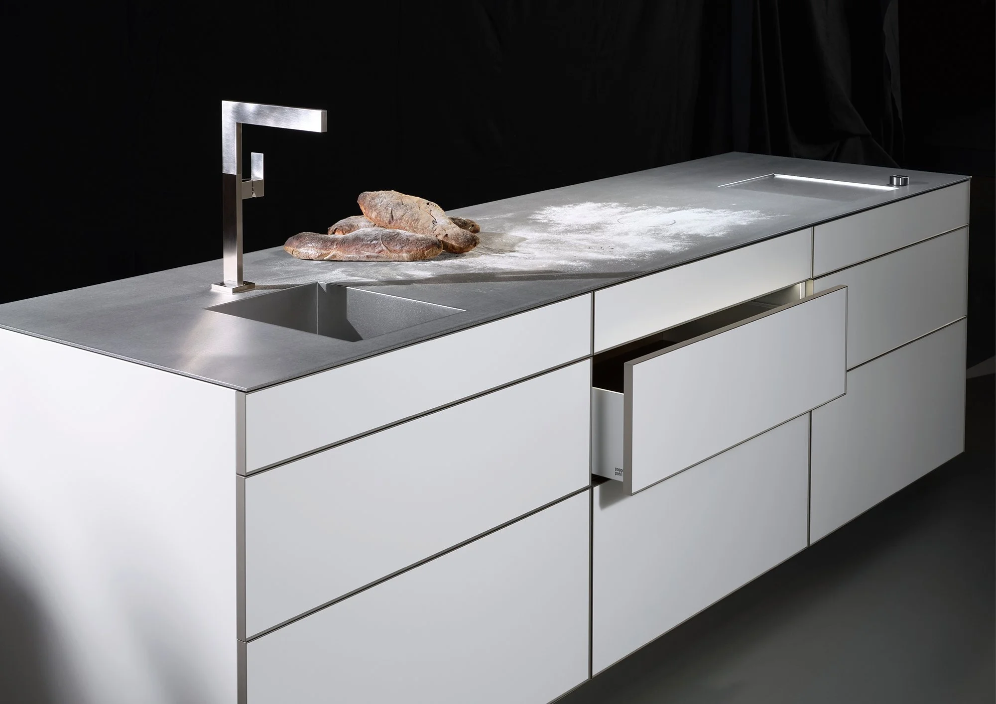 Modern kitchen countertop with a stainless steel faucet, a small pile of rocks, and flour dusted on the surface. The countertop is light-colored with multiple drawers below and a black background.