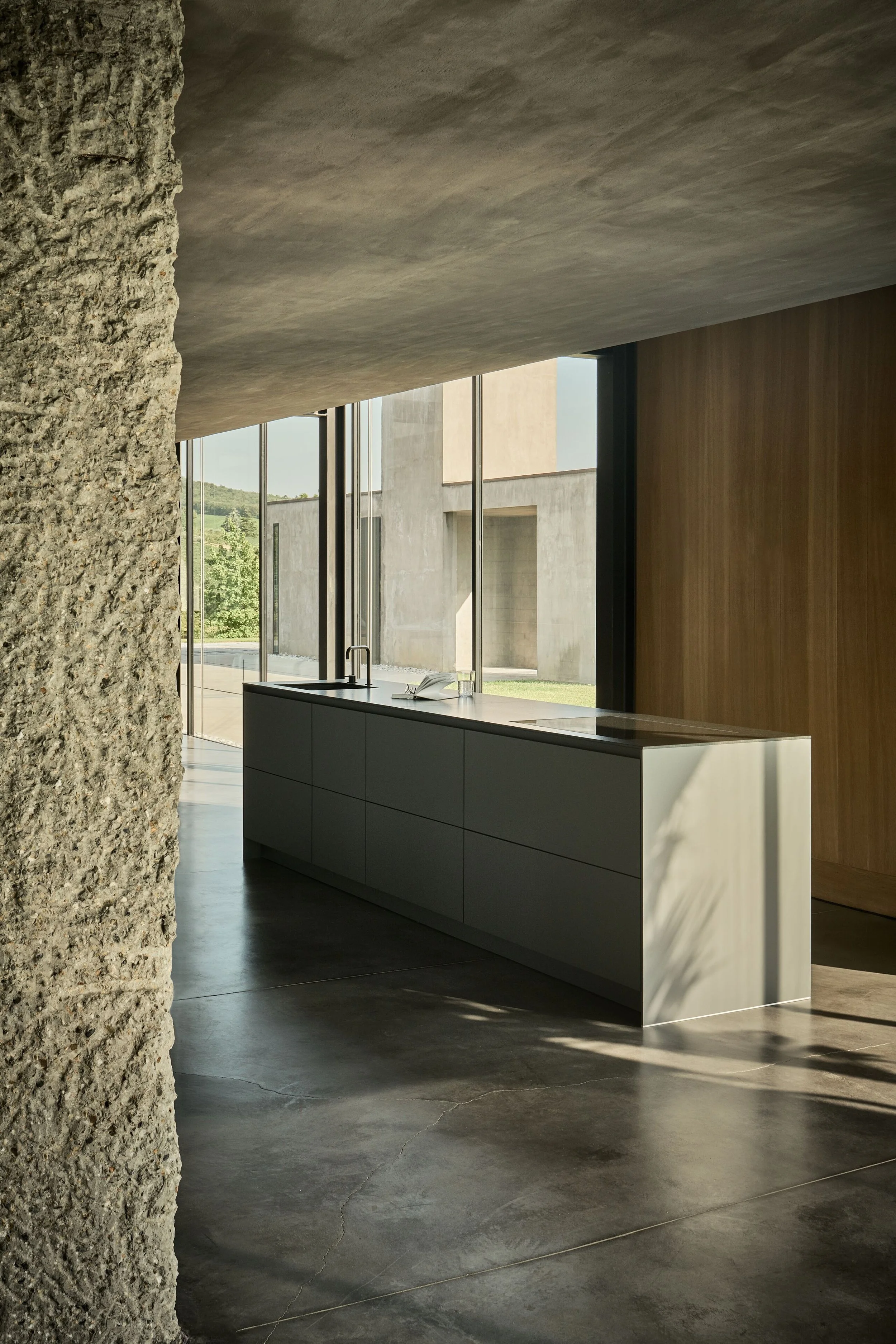 Modern interior space with textured concrete wall on the left, large windows allowing natural light, and a sleek, minimalistic kitchen island with a open book and glass on top, complemented by a wood-paneled wall.