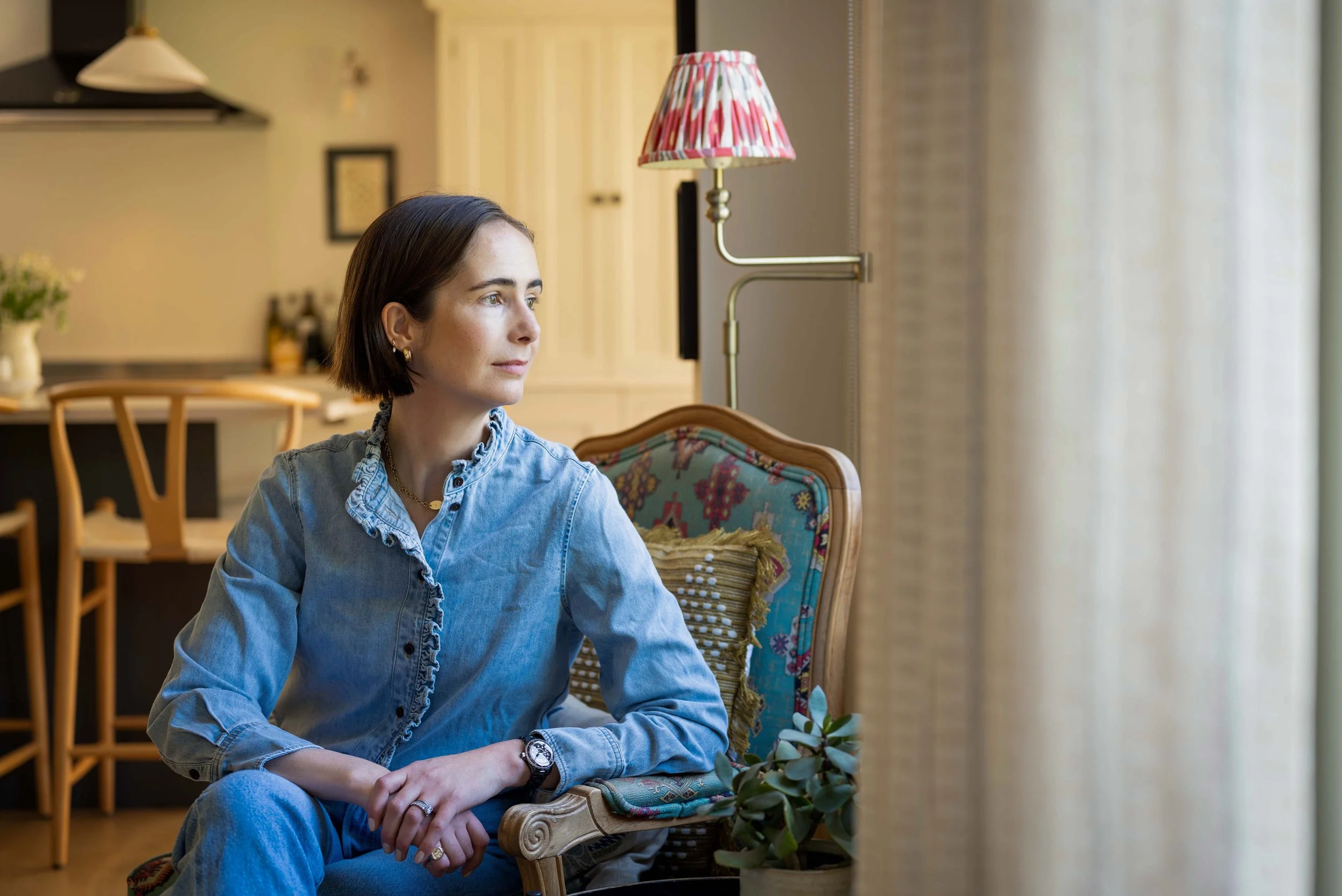 A woman with short brown hair wearing a denim shirt and watch sitting on a colorful upholstered chair in a room with warm lighting, a lamp with a pink patterned shade, and a bookshelf.