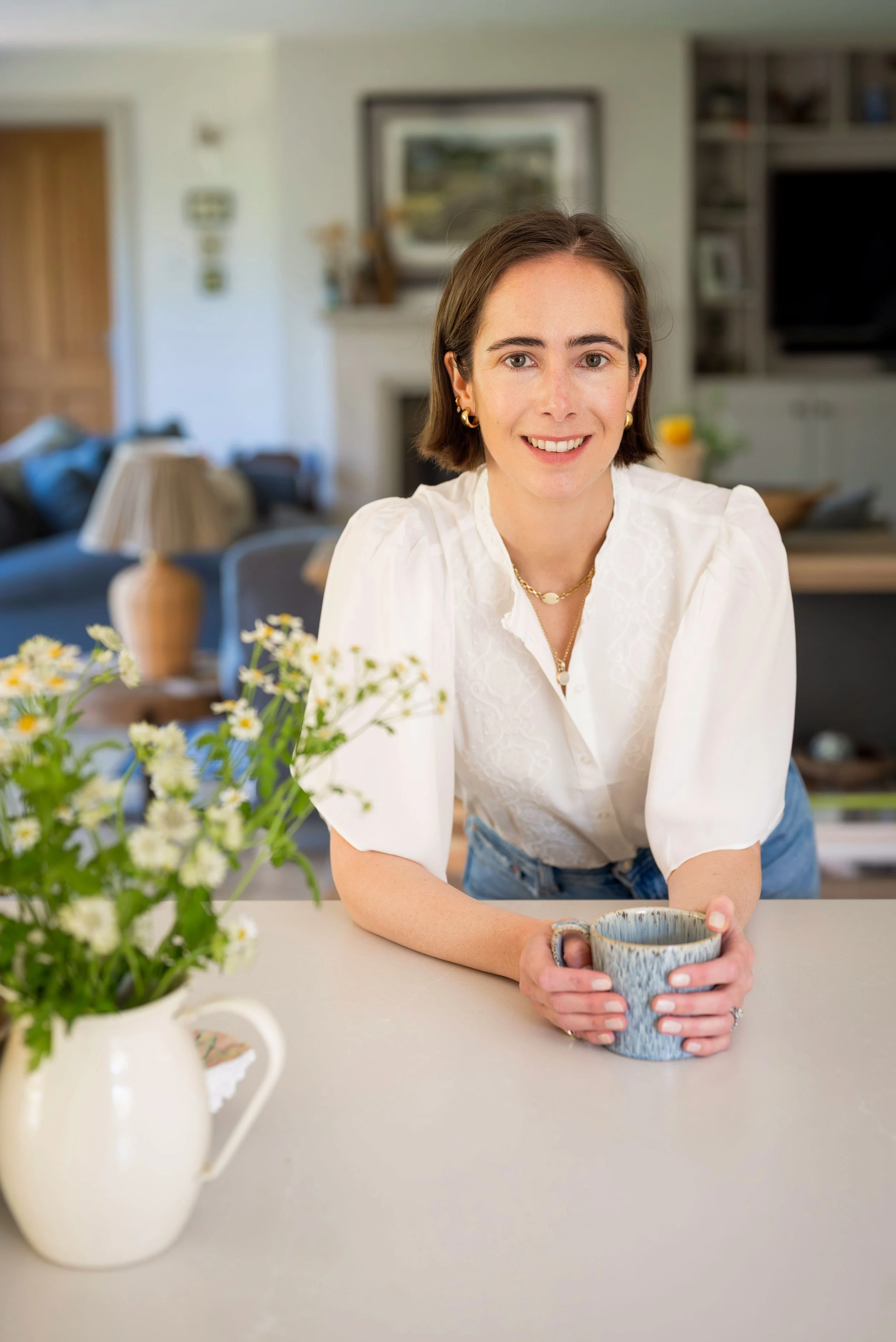 A woman with short brown hair and gold jewelry sitting at a kitchen counter, holding a ceramic mug with a textured blue and brown glaze, smiling at the camera. In the foreground, there is a white pitcher with a handle and a bouquet of small white flowers. The background shows a cozy living room with a fireplace, shelves, and a couch.
