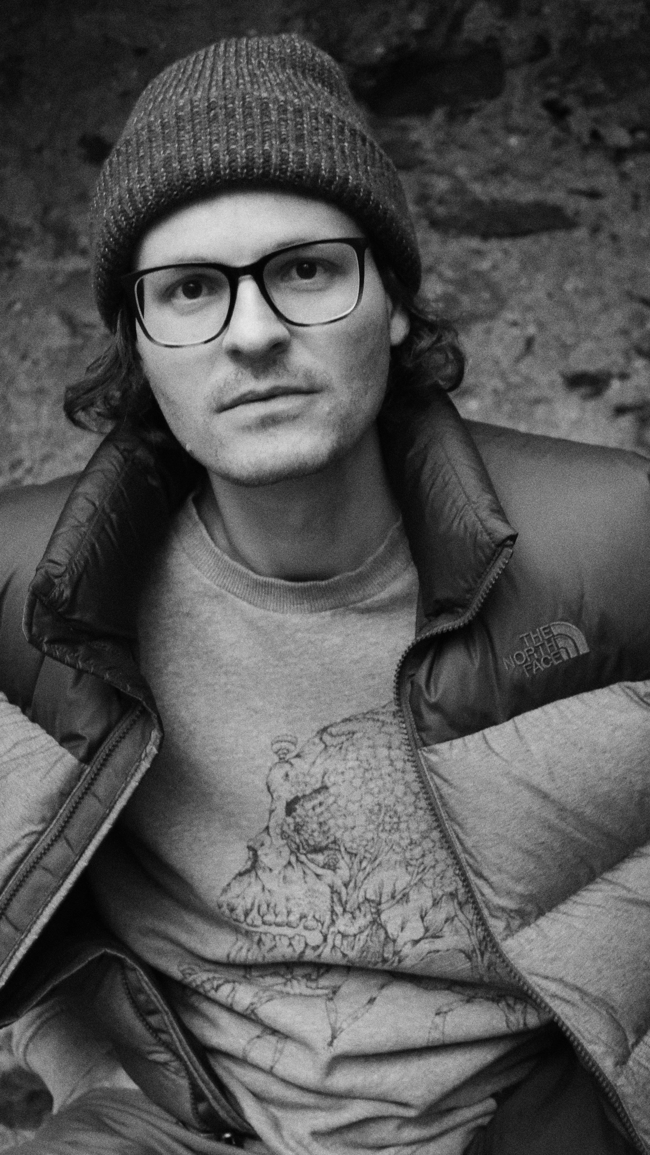 Schwarz-weiß-Foto eines Mannes mit Brille, grauem Pullover mit Tiermuster und einer North Face Jacke, trägt eine Mütze, sitzt auf einem Felsen.