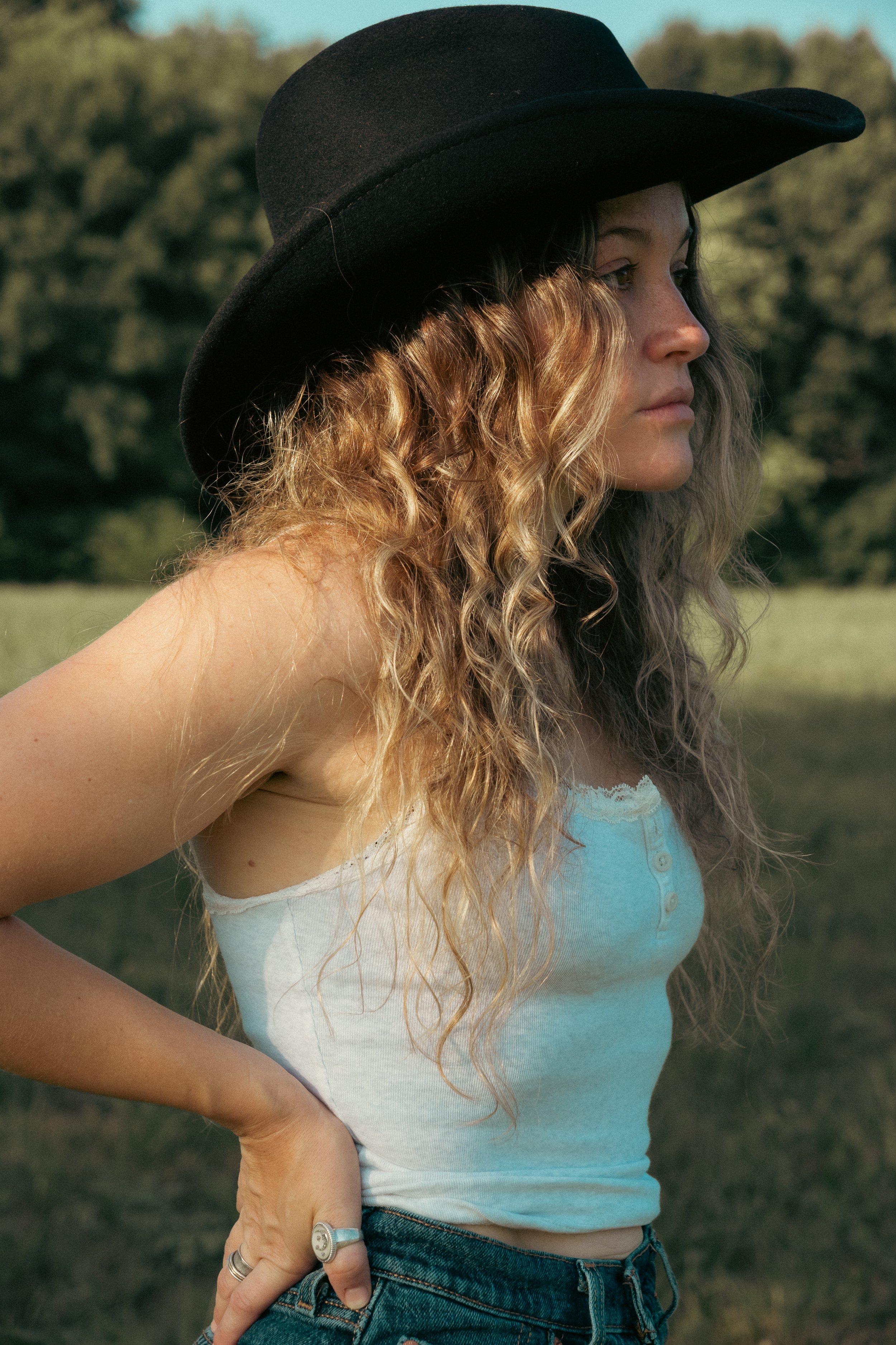 A woman with curly hair wearing a black hat and a light blue tank top stands outdoors in a field with trees in the background, looking to the right side of the image.