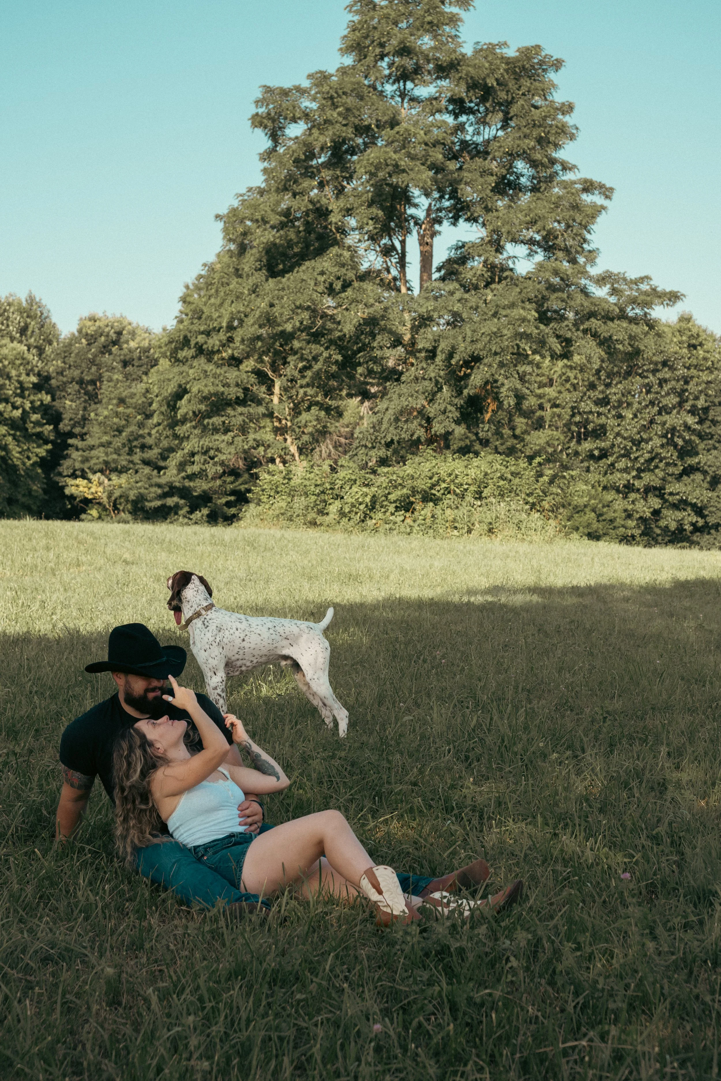 A man with a beard and black cowboy hat sitting on grass, holding a woman with long curly blonde hair and a light-colored tank top in his lap, in an open field with trees and clear sky in the background. A Dalmatian dog stands nearby.