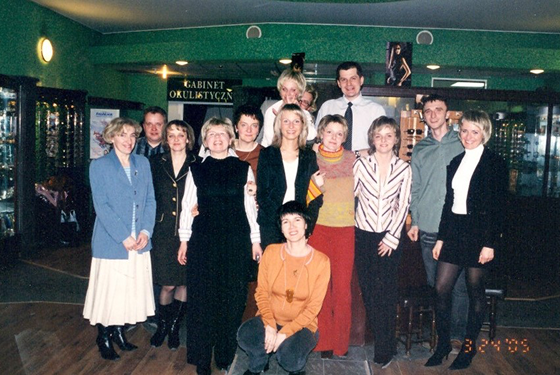 Group of ten women and three men posing for a photo in a room with dark green walls and a wooden floor, with a sign in a foreign language and some posters in the background.