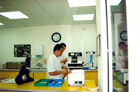 Two students in white uniforms working in a school laboratory with microscopes and scientific equipment.