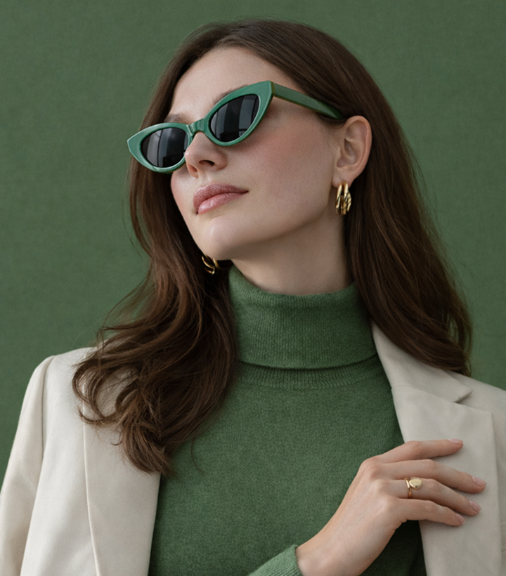 A woman with long brown hair wearing green sunglasses, gold earrings, a green turtleneck, and a cream blazer against a green background.