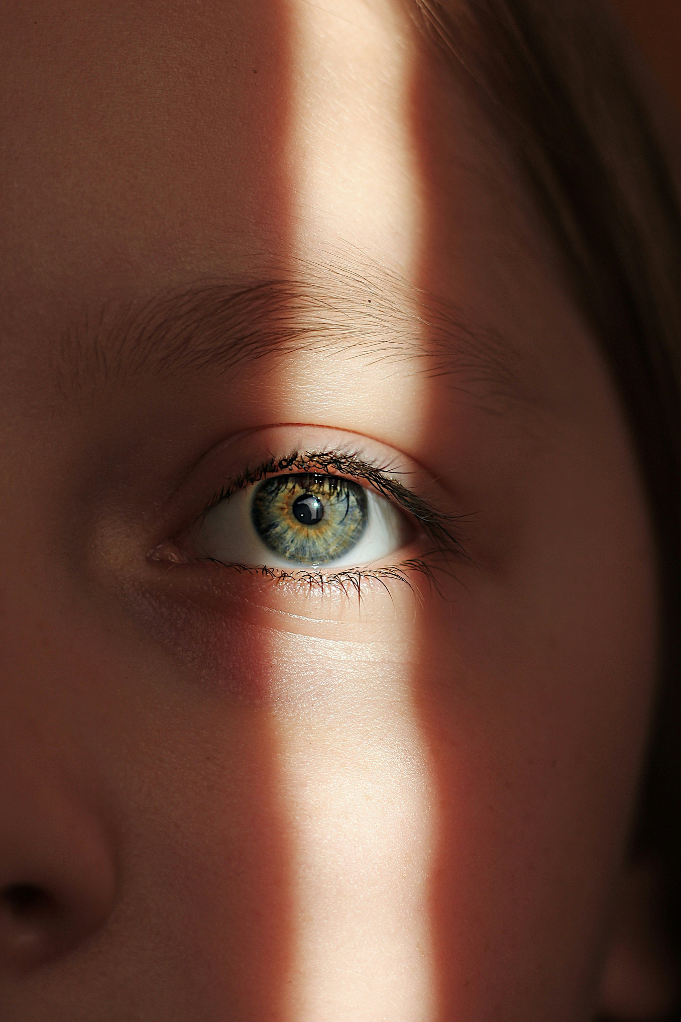Close-up of a human eye with blue and green iris, partially covered by eyelids and eyelashes, with sunlight casting a shadow across the face.
