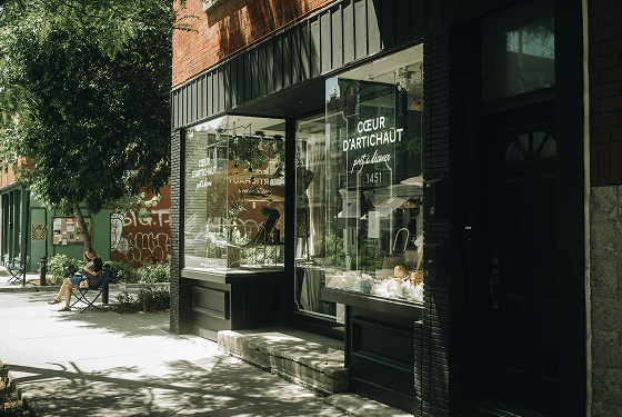 A storefront with large glass windows and a black awning, displaying jewelry inside. A woman sits on a bench outside in front of a tree, with graffiti on a wall nearby.