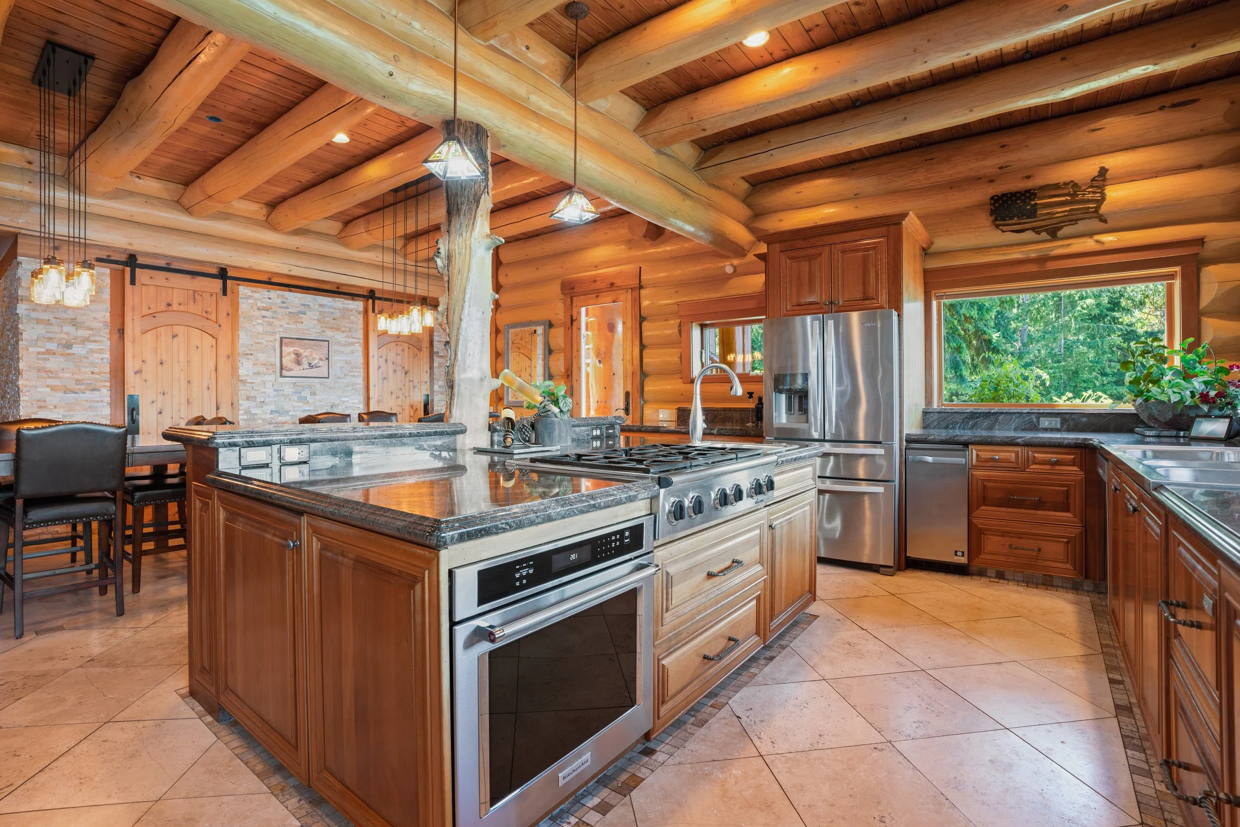 Interior view of a log cabin kitchen with wooden beams on the ceiling, a large window showing greenery outside, and kitchen appliances including a stainless steel refrigerator and oven, with a central island and seating area.