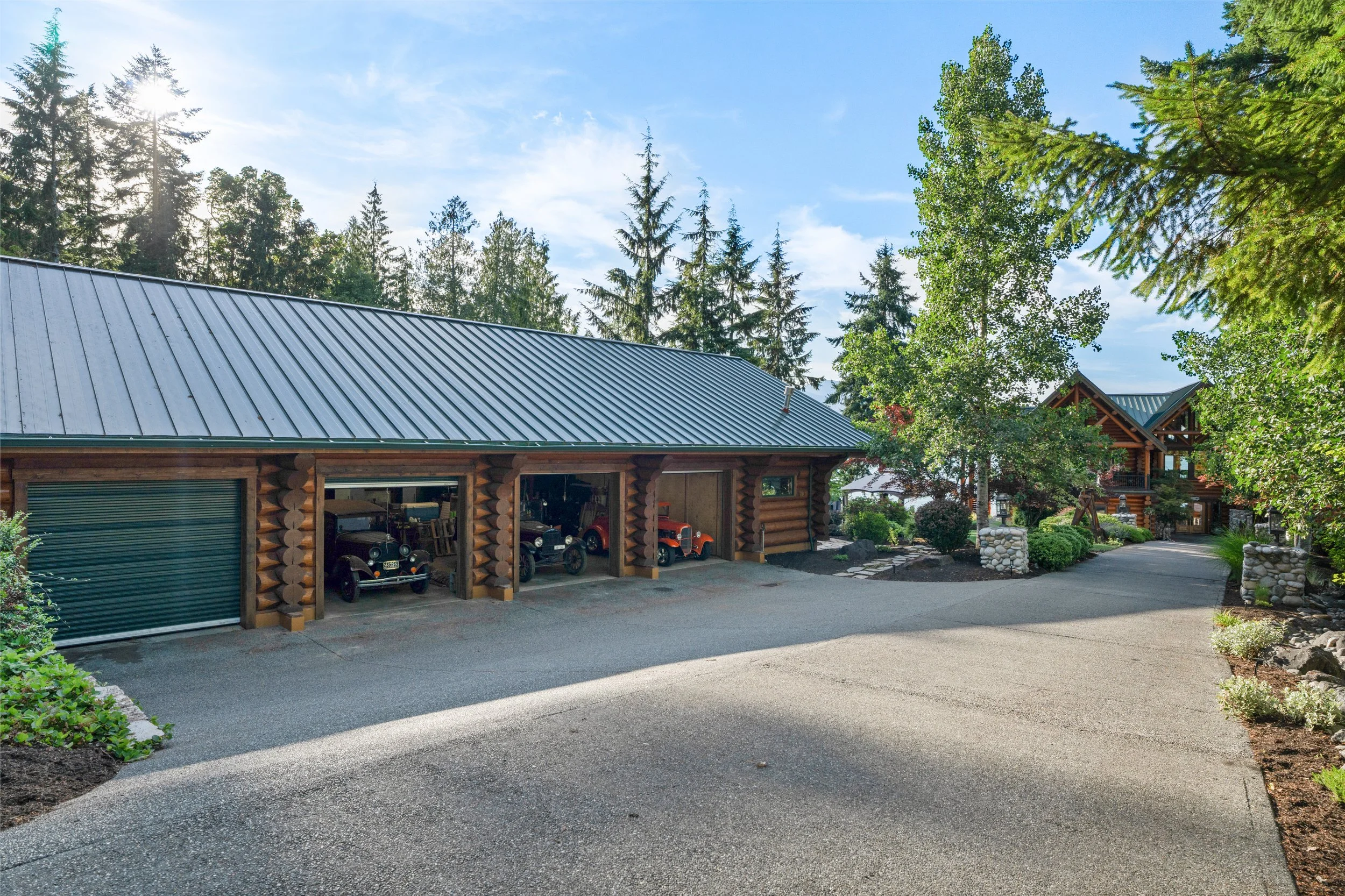 View of a wooden garage with a metal roof, cars inside, surrounded by trees and greenery in a mountainous area under a blue sky.