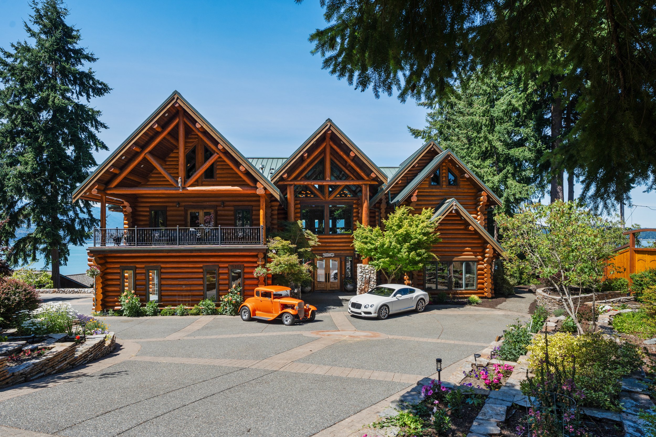 A large wooden log cabin-style house with a green metal roof, surrounded by tall evergreen trees and a landscaped garden with colorful flowers. In front, there are two cars parked: a vintage orange car and a modern white sports car. The sky is clear and blue.