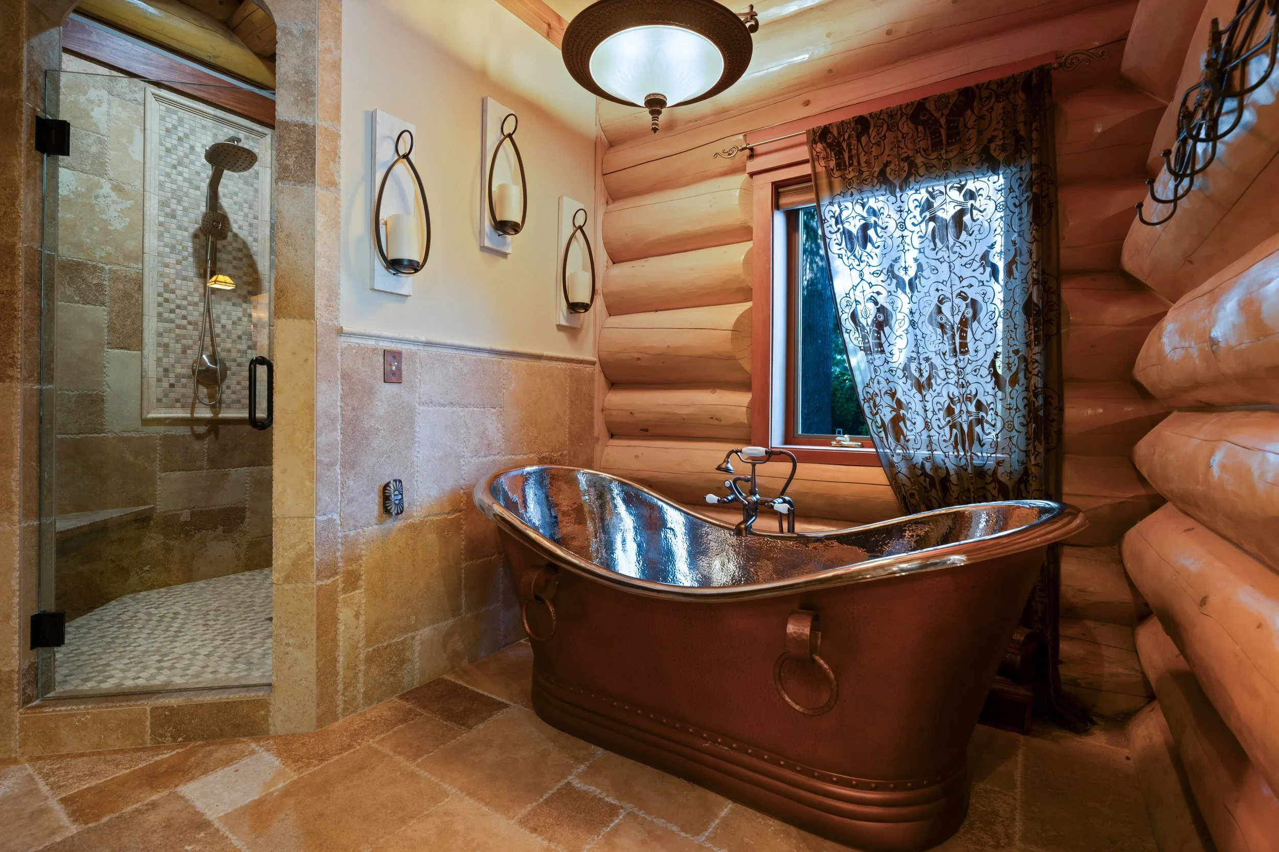 A rustic log cabin bathroom featuring a copper clawfoot tub, a shower with a glass door, stone tile flooring, log walls, a window with patterned curtains, and wall-mounted candle holders.