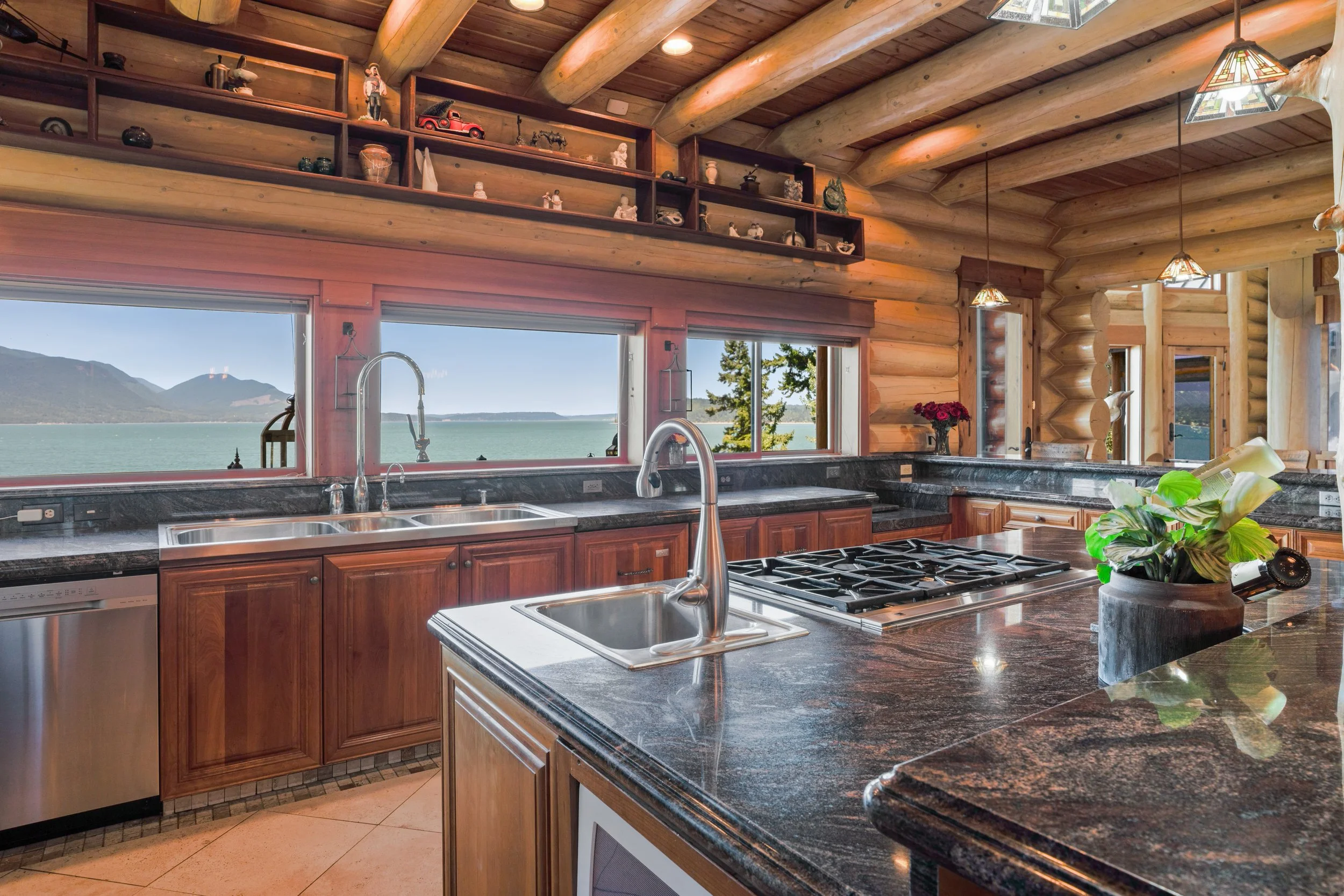Log cabin kitchen with large windows showing a scenic mountain and lake view, wooden log walls, granite countertops, stainless steel sink, stove, hanging lamps, decorative shelves with ornaments, and potted plant.