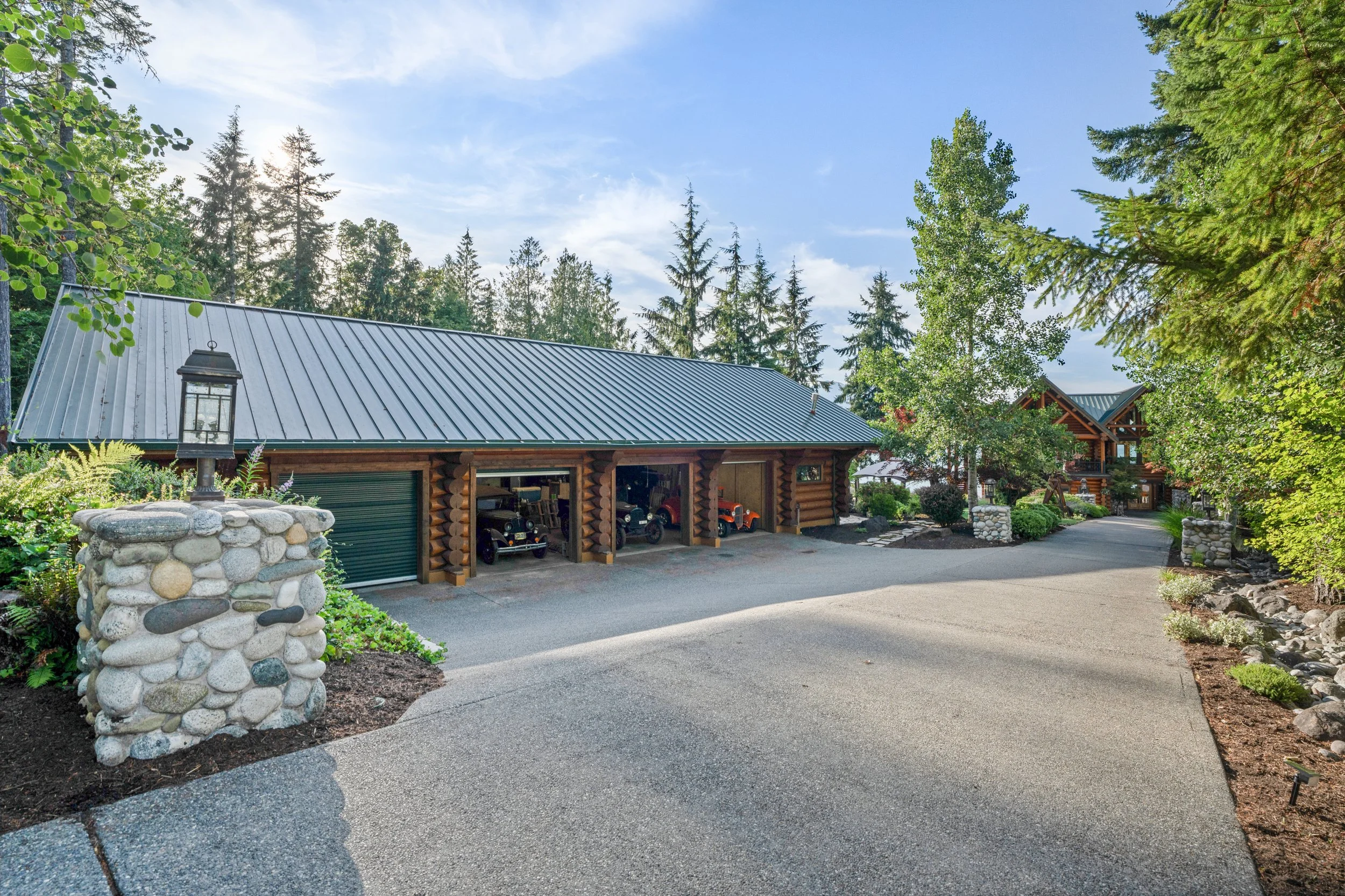 A spacious driveway leading to a log cabin-style house with a metal roof, surrounded by lush trees and landscaped with plants and stone accents.