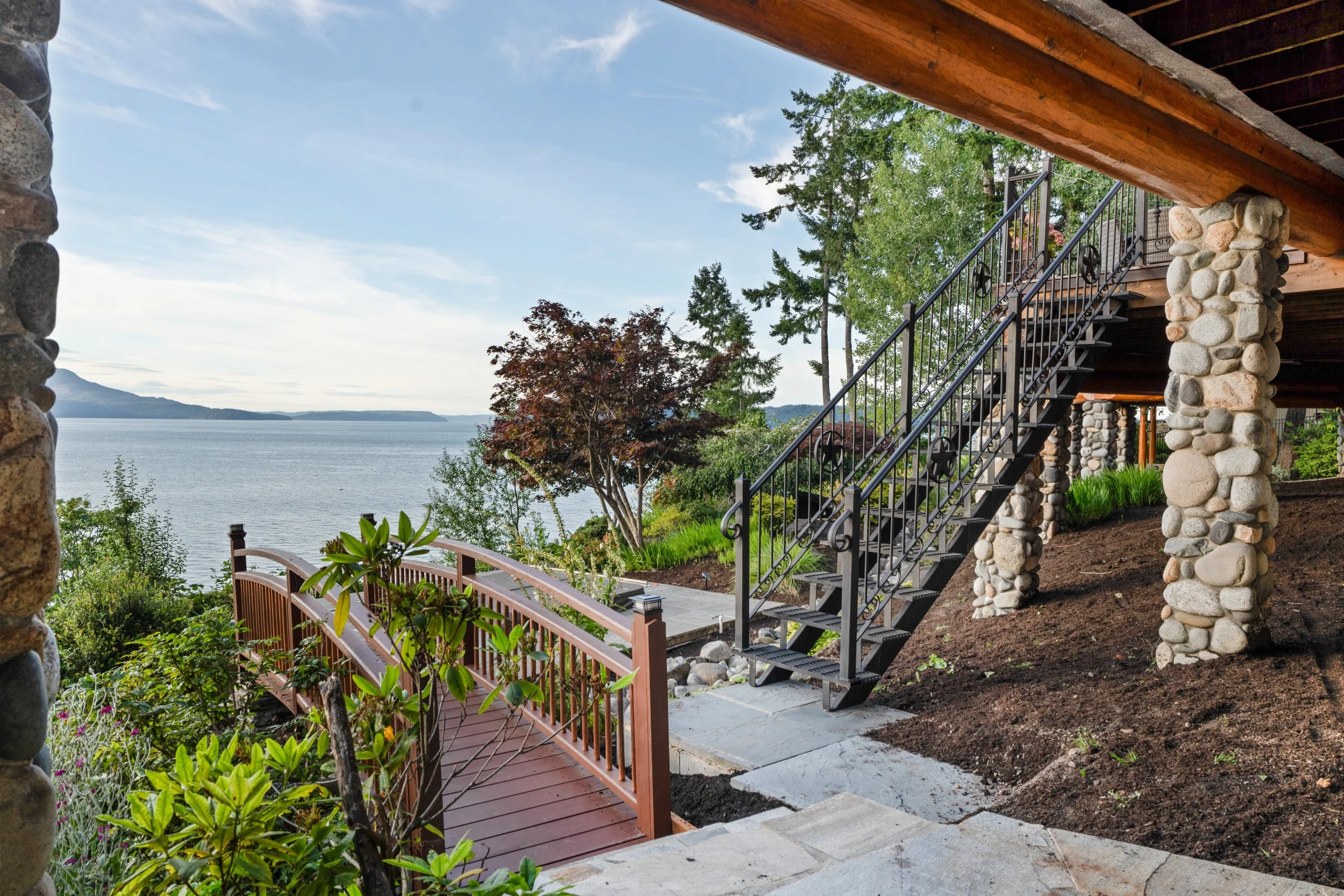View of a lakeside yard from under a wooden deck with stone columns, metal staircase, green trees, and a body of water with distant mountains.