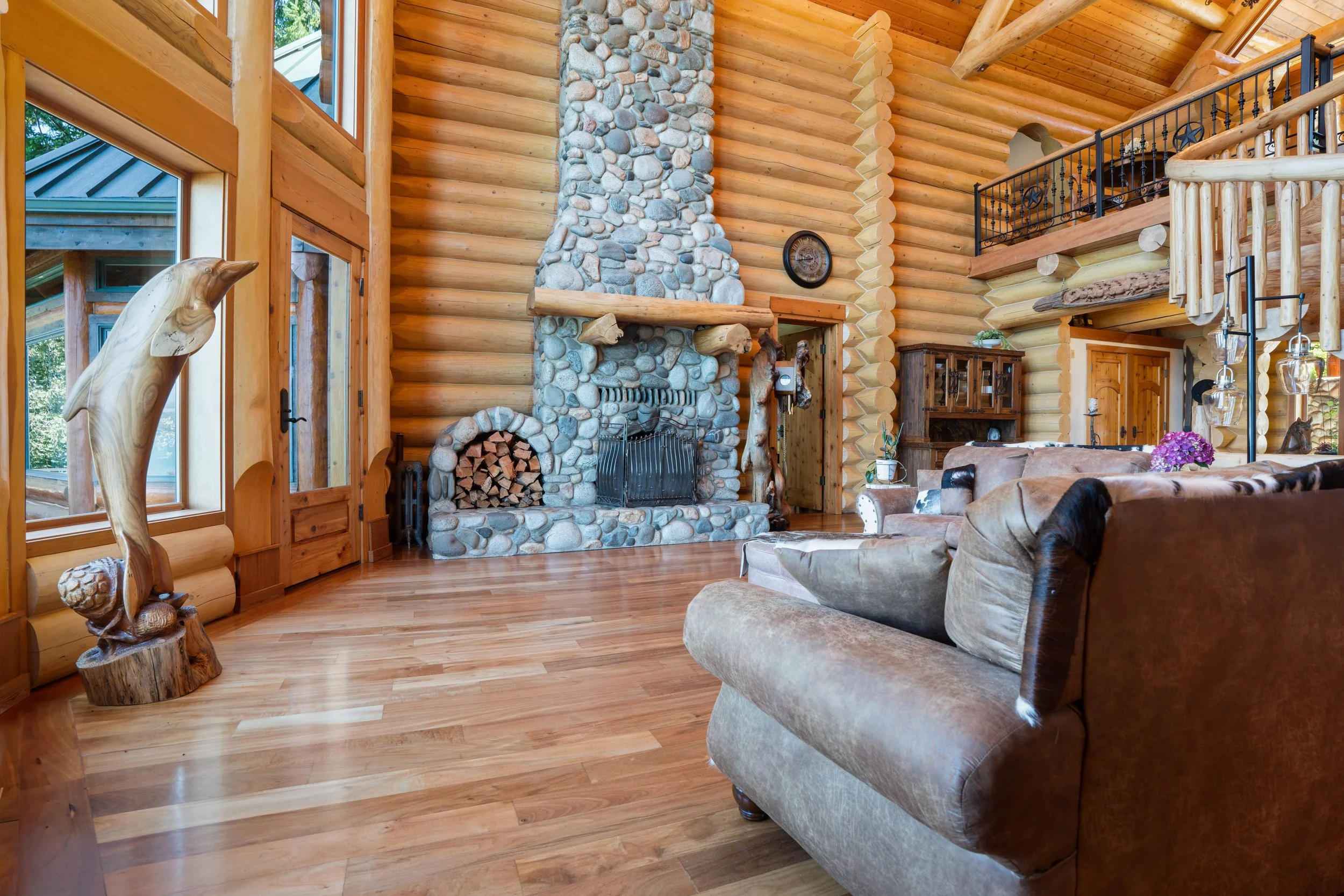 Interior view of a log cabin living room with a stone fireplace, wooden log walls, a leather sofa, and decorative wood and animal sculptures.