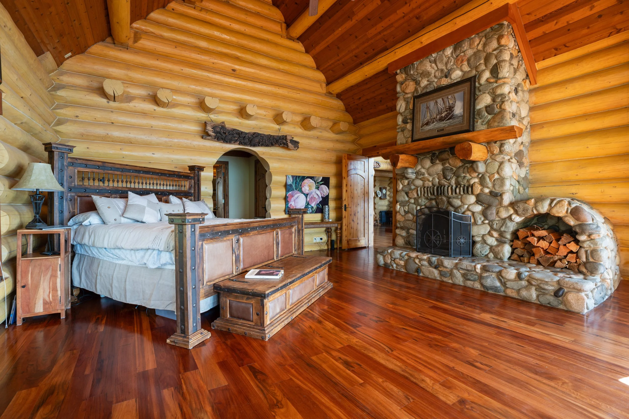 A cozy rustic bedroom with wooden log walls and ceiling, a stone fireplace with a wooden mantel, a framed picture of a ship, a wooden bed with white bedding, a nightstand with a lamp, and hardwood flooring.