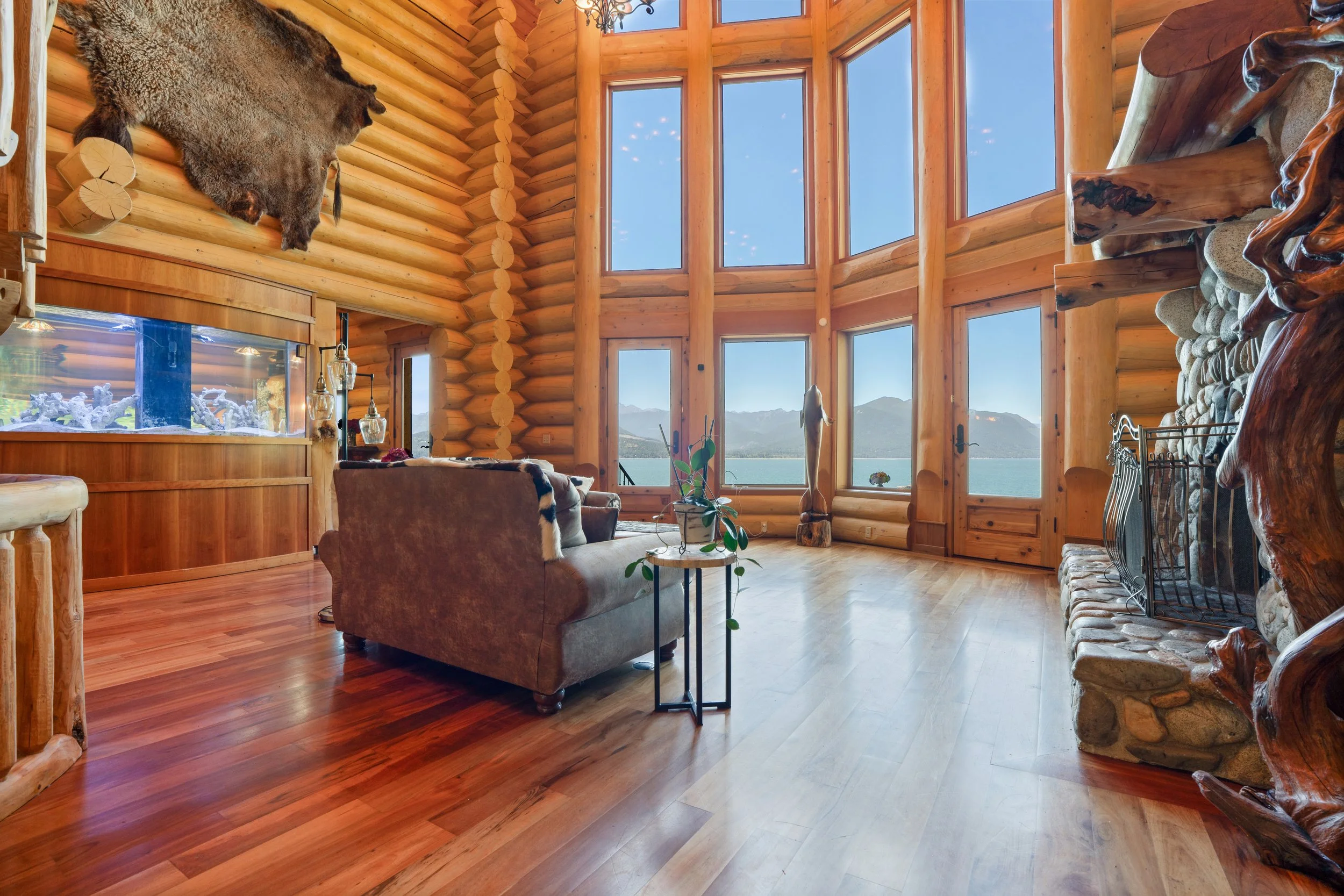 Interior of a log cabin living room with large windows overlooking mountains and water, featuring a mounted animal hide on the wall, a stone fireplace, wooden furniture, and rustic decor.