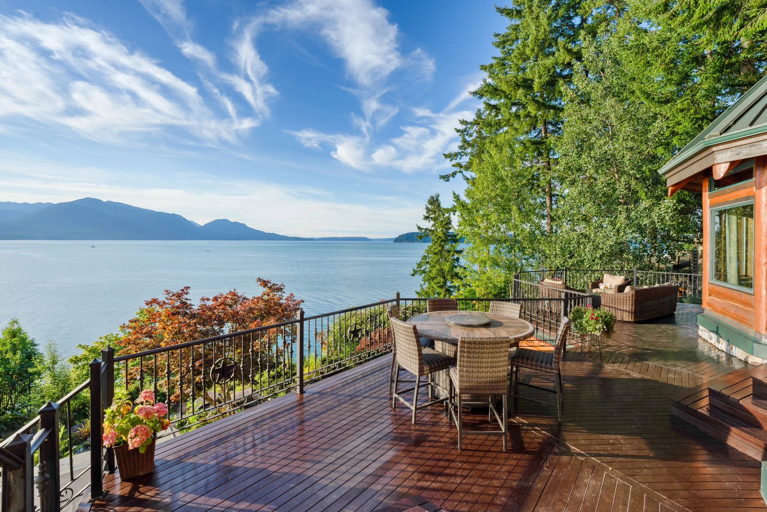 A spacious wooden deck overlooking a body of water with mountains in the distance. The deck has a round table and several wicker chairs, with potted flowers and lush green trees providing shade and scenery. Part of a house with large windows and a green roof is visible on the right side.