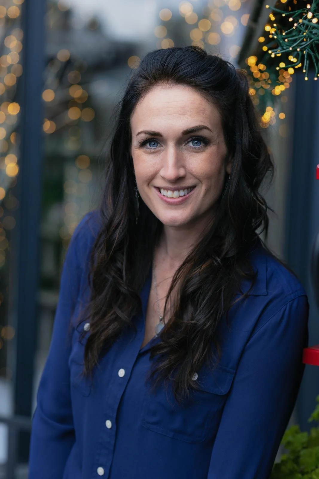 A woman with long dark hair and blue eyes smiling in front of a blurred background with Christmas lights and decorations.