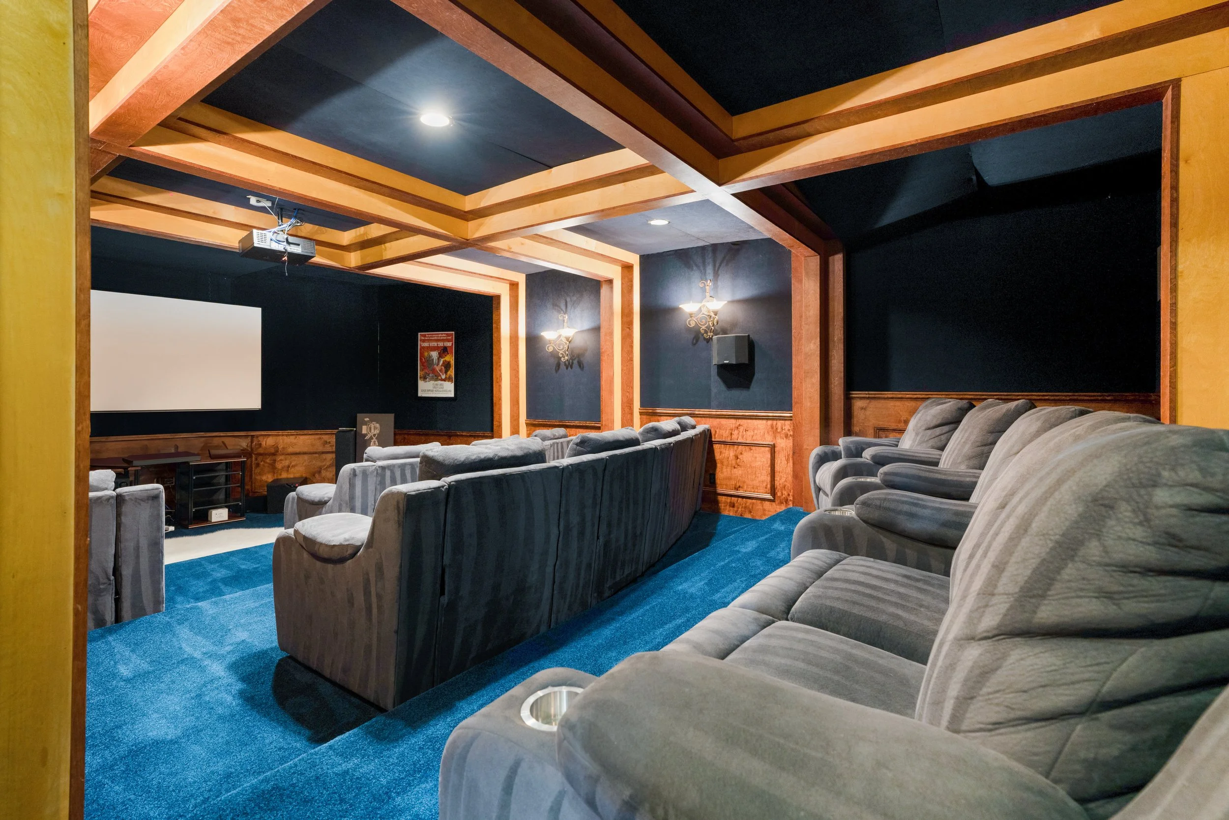 A cozy home theater room with a large screen, multiple gray plush armchairs, wall-mounted sconces, and a blue carpet, with a ceiling decorated with wooden trim and blue panels.