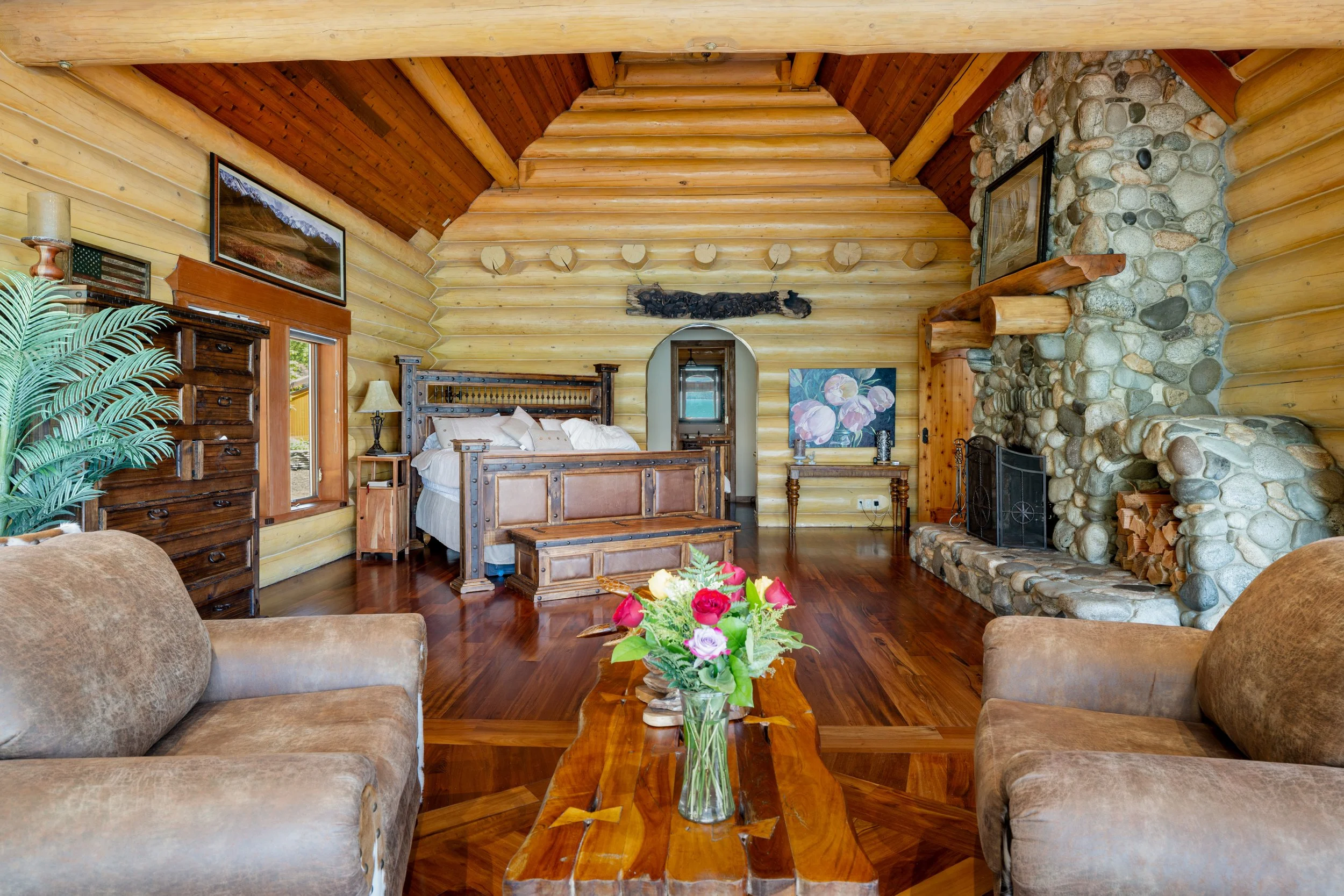 Cozy living room with wood logs and stone fireplace, wooden furniture, and floral decor.
